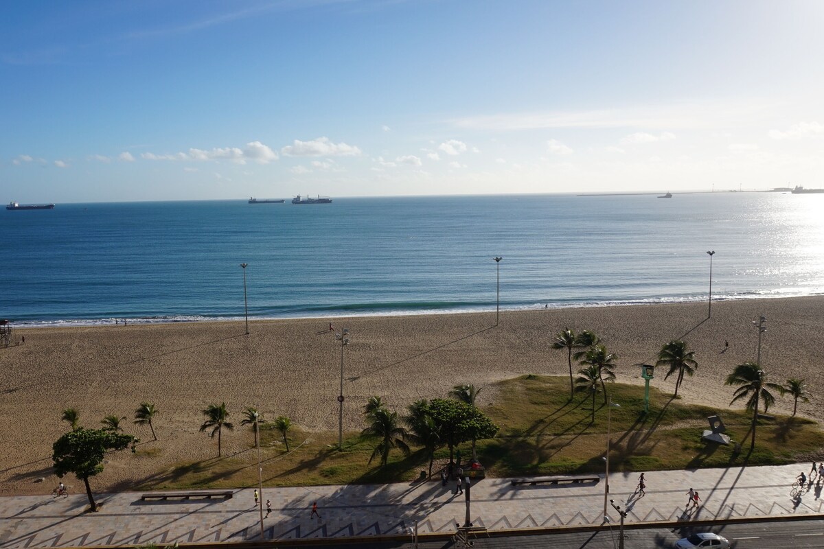 A beach with a paved walkway, palm trees, and a view of the ocean with ships in the distance.