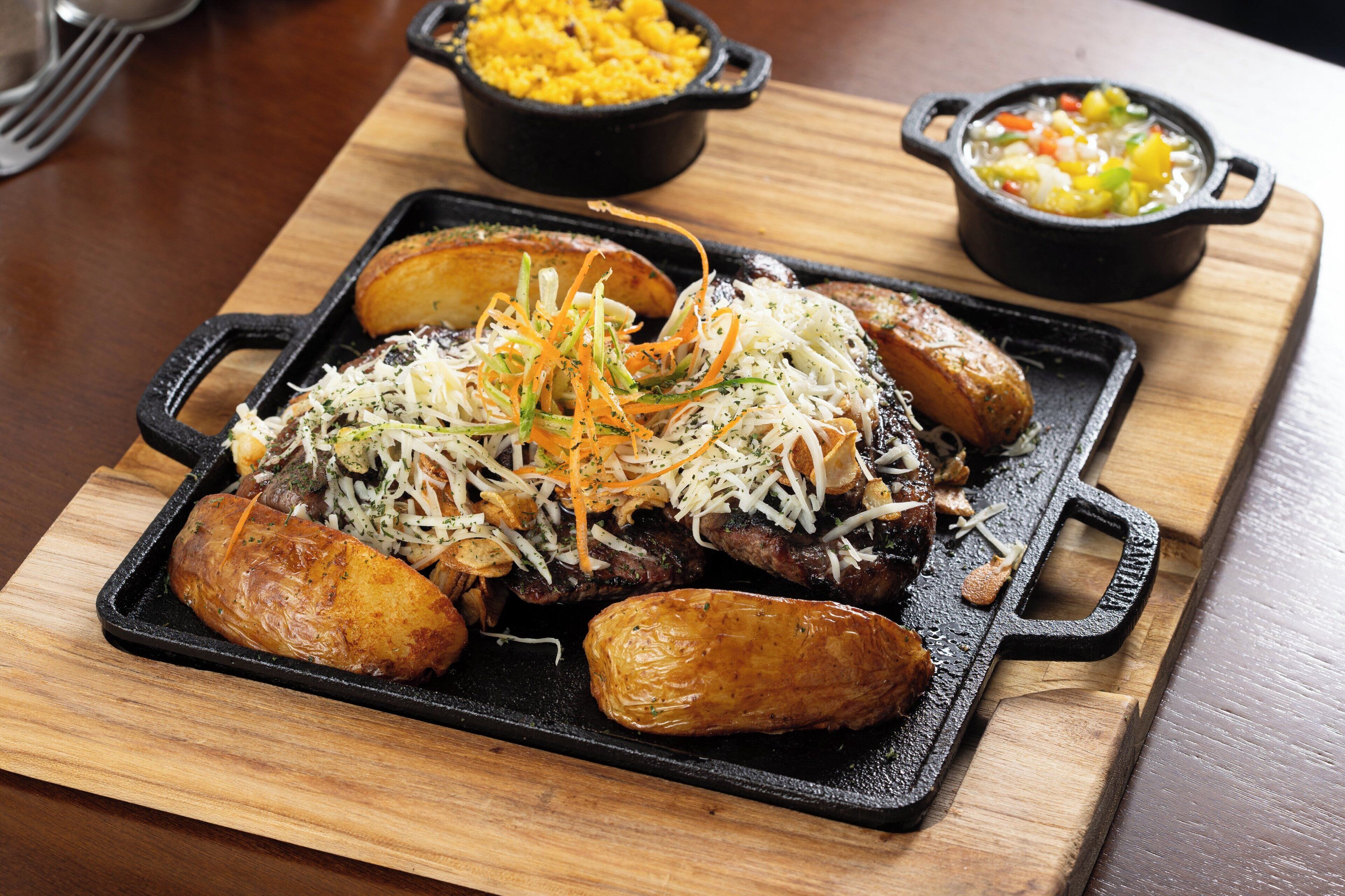 Una sartén de hierro fundido con papas asadas, carne y queso rallado sobre una mesa de madera.