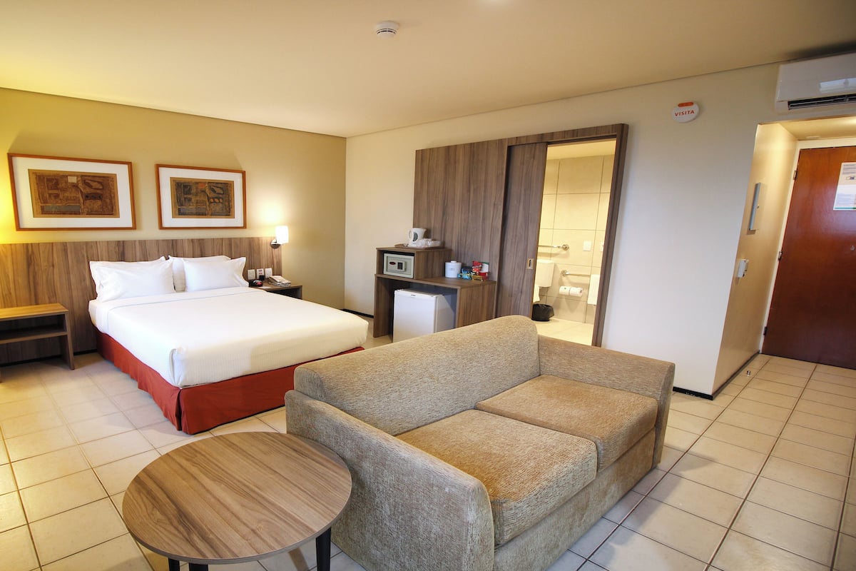 Quarto de hotel com cama, sofá, pequena mesa, mesa de cabeceira, frigorífico e casa de banho visível através de uma porta aberta, com um sinal 'VISITA' na parede.
