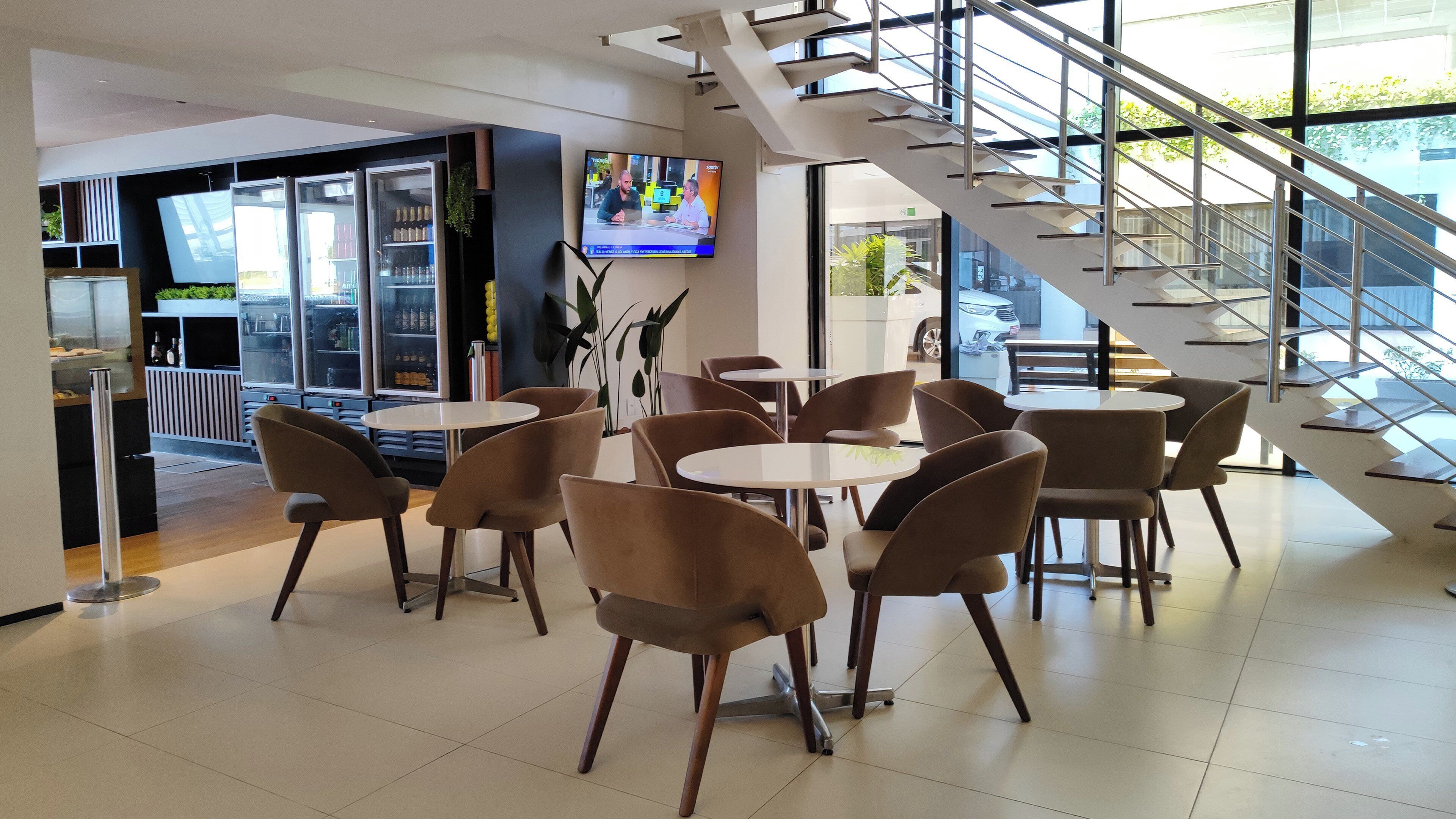 Un espacio interior moderno con mesas y sillas redondas, una escalera y una pantalla de televisión mostrando un noticiero.
