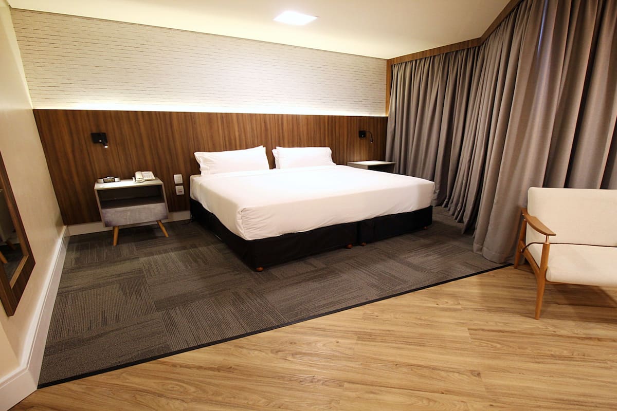 Quarto de hotel com cama, escrivaninha, cadeira, mesa de cabeceira e luminária fixada na parede.