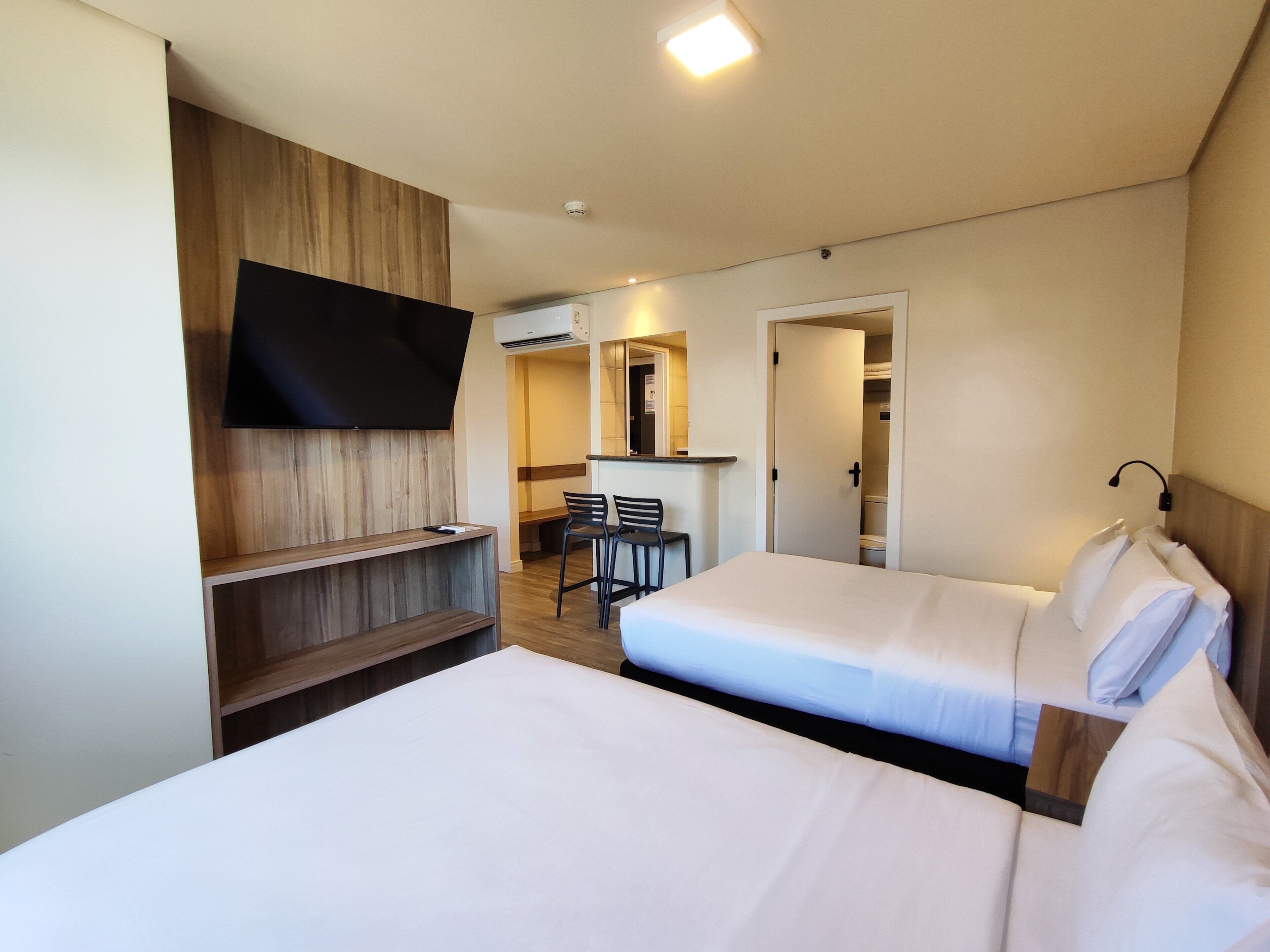 A hotel room with two beds, a desk, a chair, a TV mounted on the wall, and an air conditioning unit.