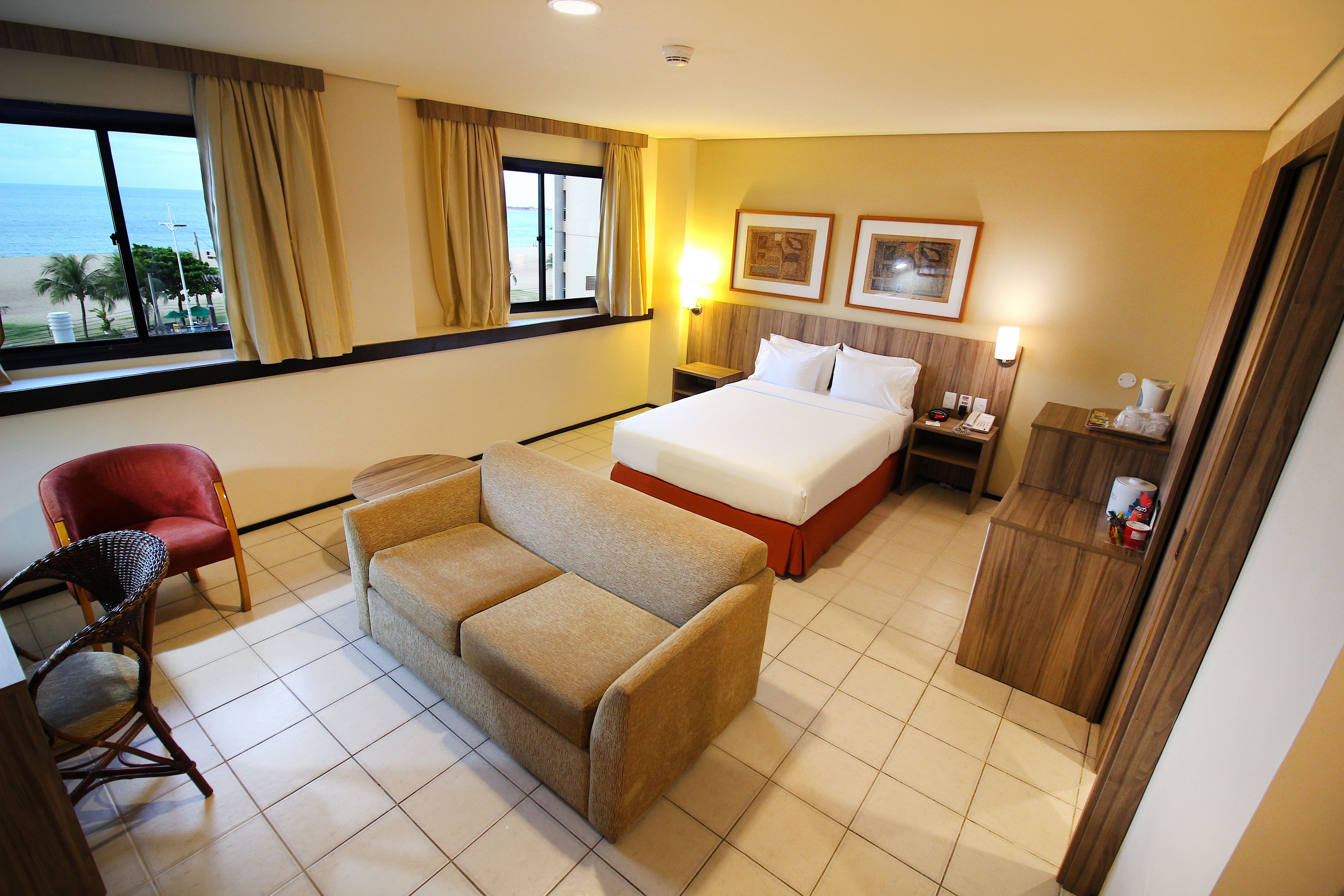 Ein Hotelzimmer mit Bett, einer Couch, einem Sessel, einem Nachttisch und Blick auf den Strand.