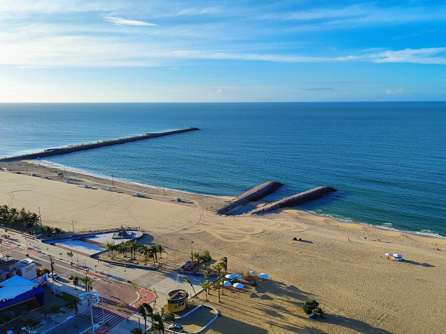A beach with a long pier extending into the sea, a sandy beach with umbrellas, and a road leading to a building with a blue roof.