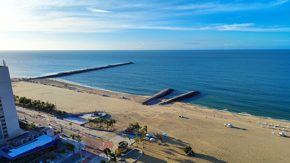 A beach with a long pier extending into the sea, a sandy beach with umbrellas, and a road leading to a building with a blue roof.