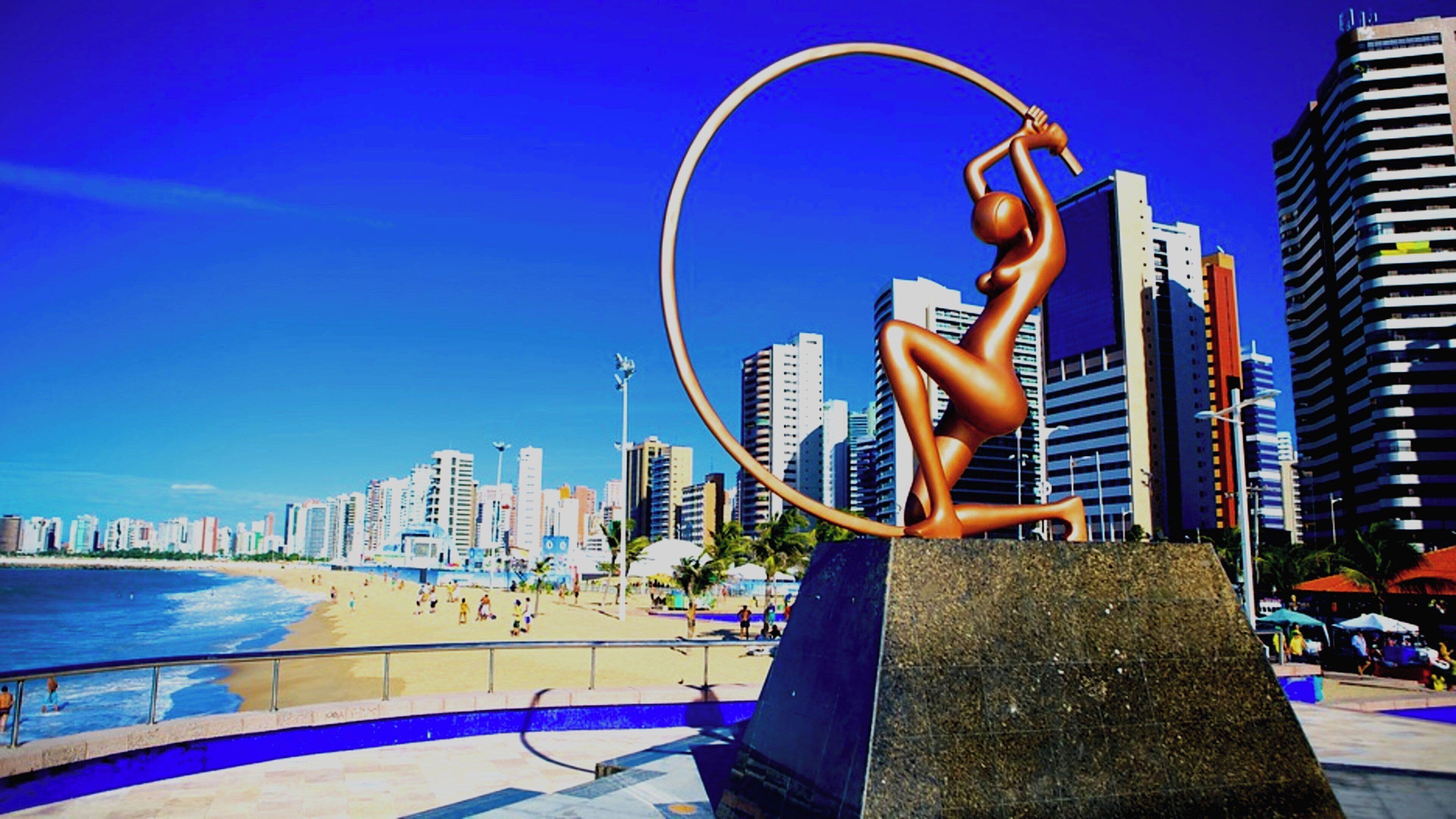 Une sculpture représentant une figure humaine effectuant un numéro avec un cerceau, le tout se détachant sur un arrière-plan composé d’une plage et de la silhouette d’une ville.