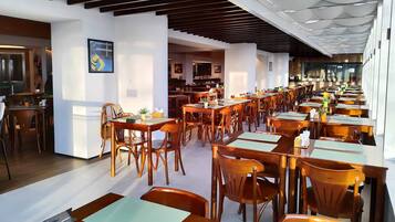 A spacious restaurant with wooden tables and chairs, a modern ceiling design, and framed artwork on the walls.