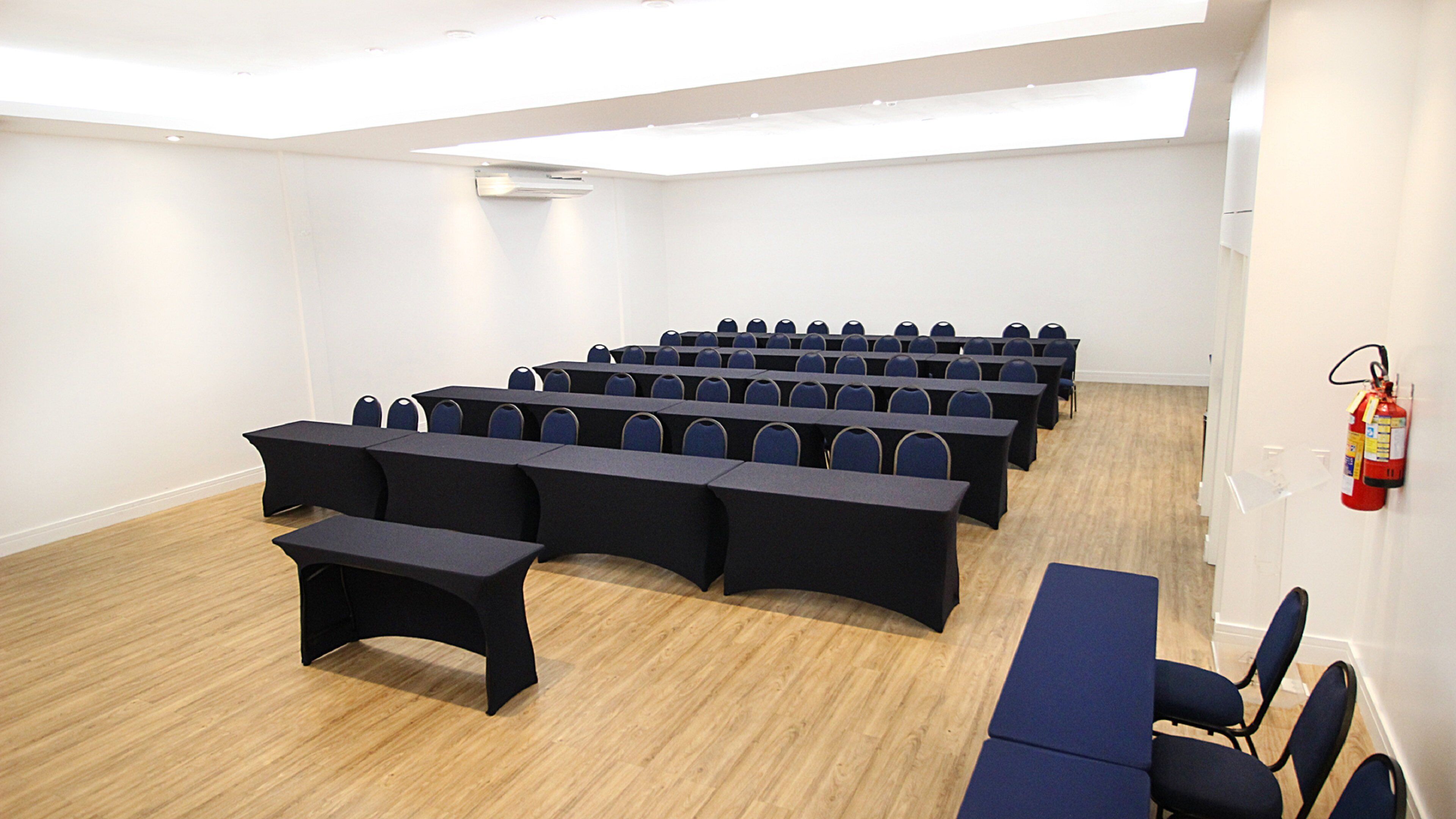 Un salón de conferencias con filas de mesas y sillas, un extintor en la pared y suelo de madera.