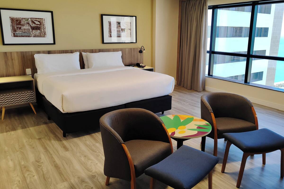 Quarto de hotel com cama, duas mesas de cabeceira, escrivaninha, cadeira, mesa pequena e uma grande janela com vista para o mar.