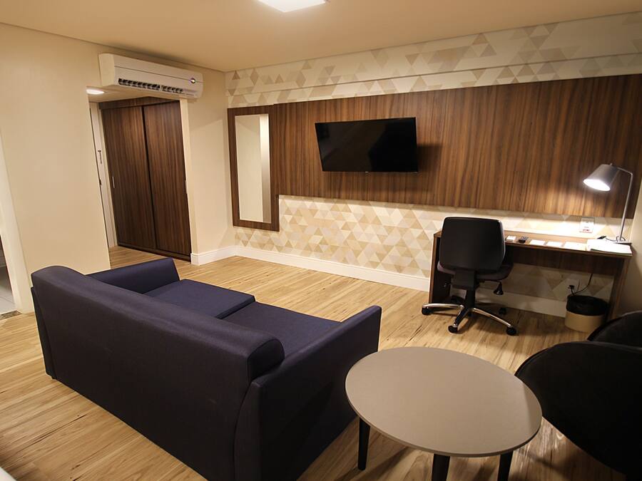 A modern living room with a sofa, a small round table, and a desk with a chair. There is a flat-screen TV mounted on the wall, a window with blinds, and a kitchen area visible in the background.