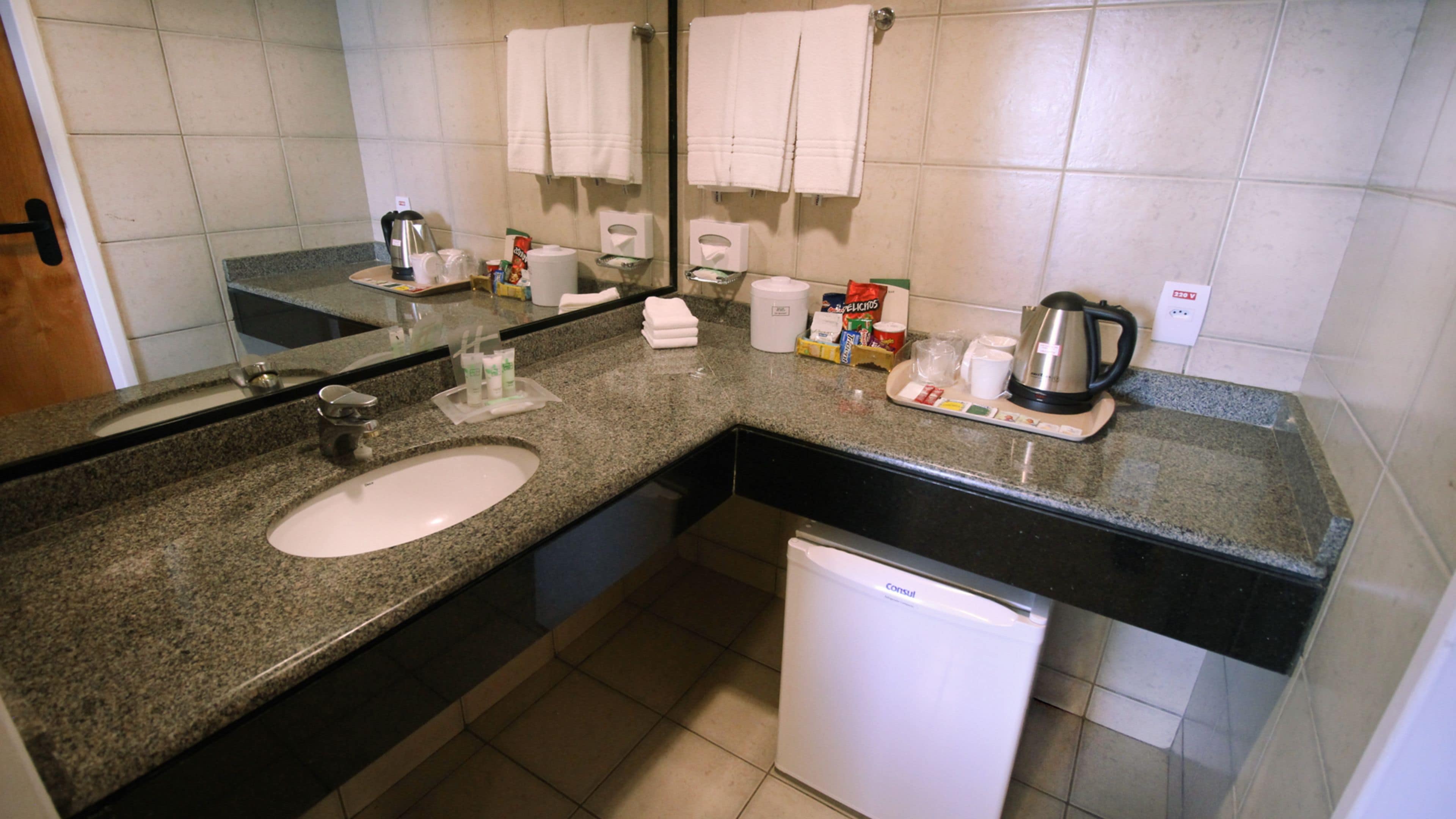 Un baño con lavabo, espejo, toallero, cafetera y un refrigerador blanco pequeño.
