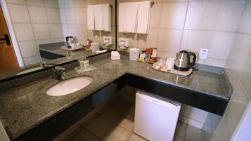 Un baño con lavabo, espejo, toallero, cafetera y un refrigerador blanco pequeño.