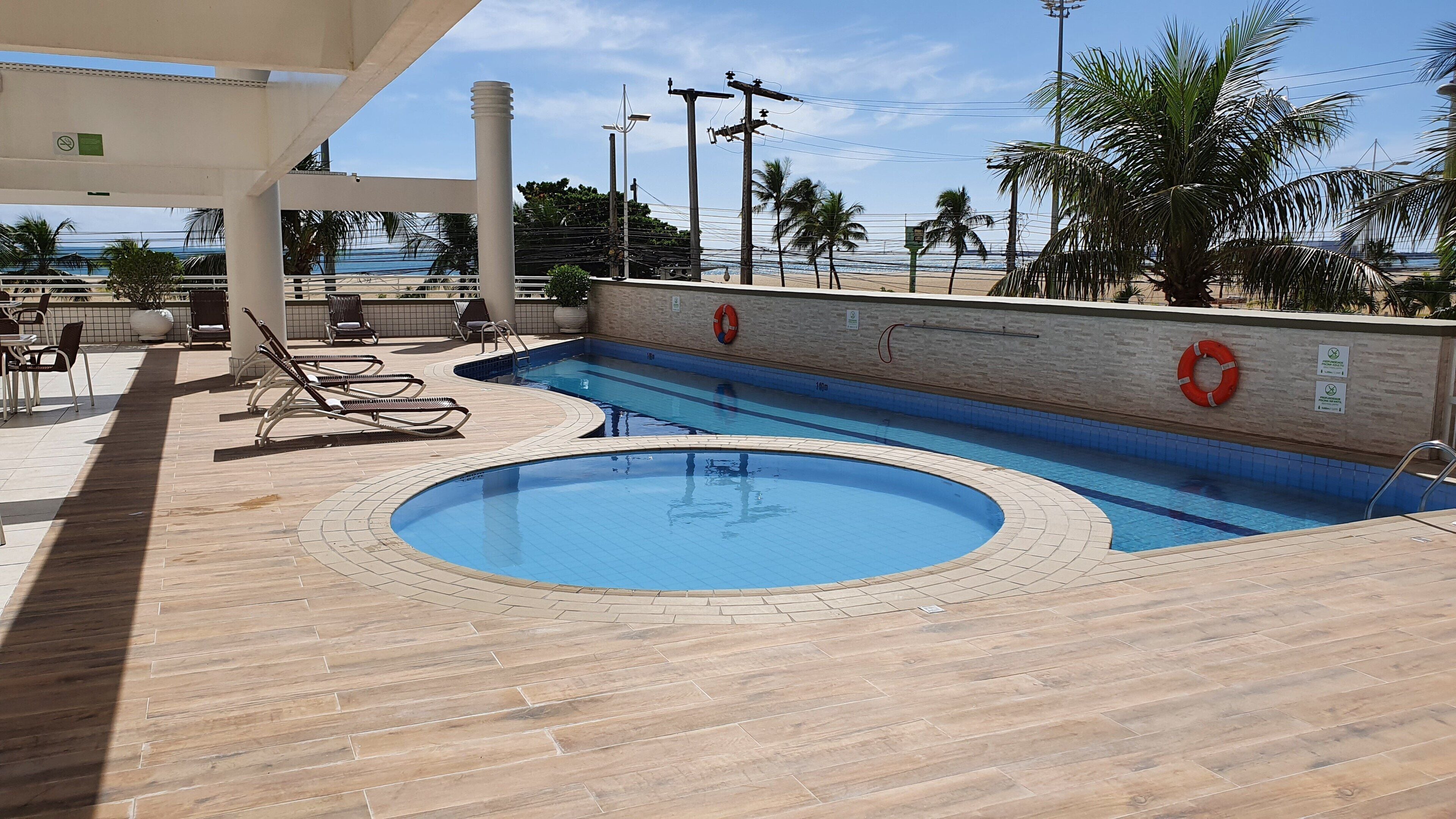 A swimming pool with lounge chairs and lifebuoys, surrounded by palm trees and a view of the ocean.