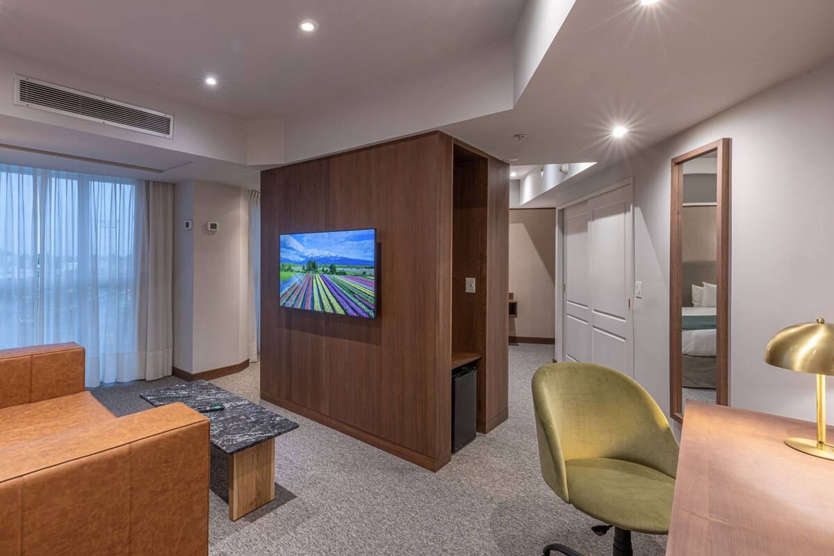 Habitación de hotel con un televisor de pantalla plana grande montado en un panel de madera, un sofá de cuero marrón, un sillón verde, una pequeña mesa de centro de madera y un escritorio con una lámpara.