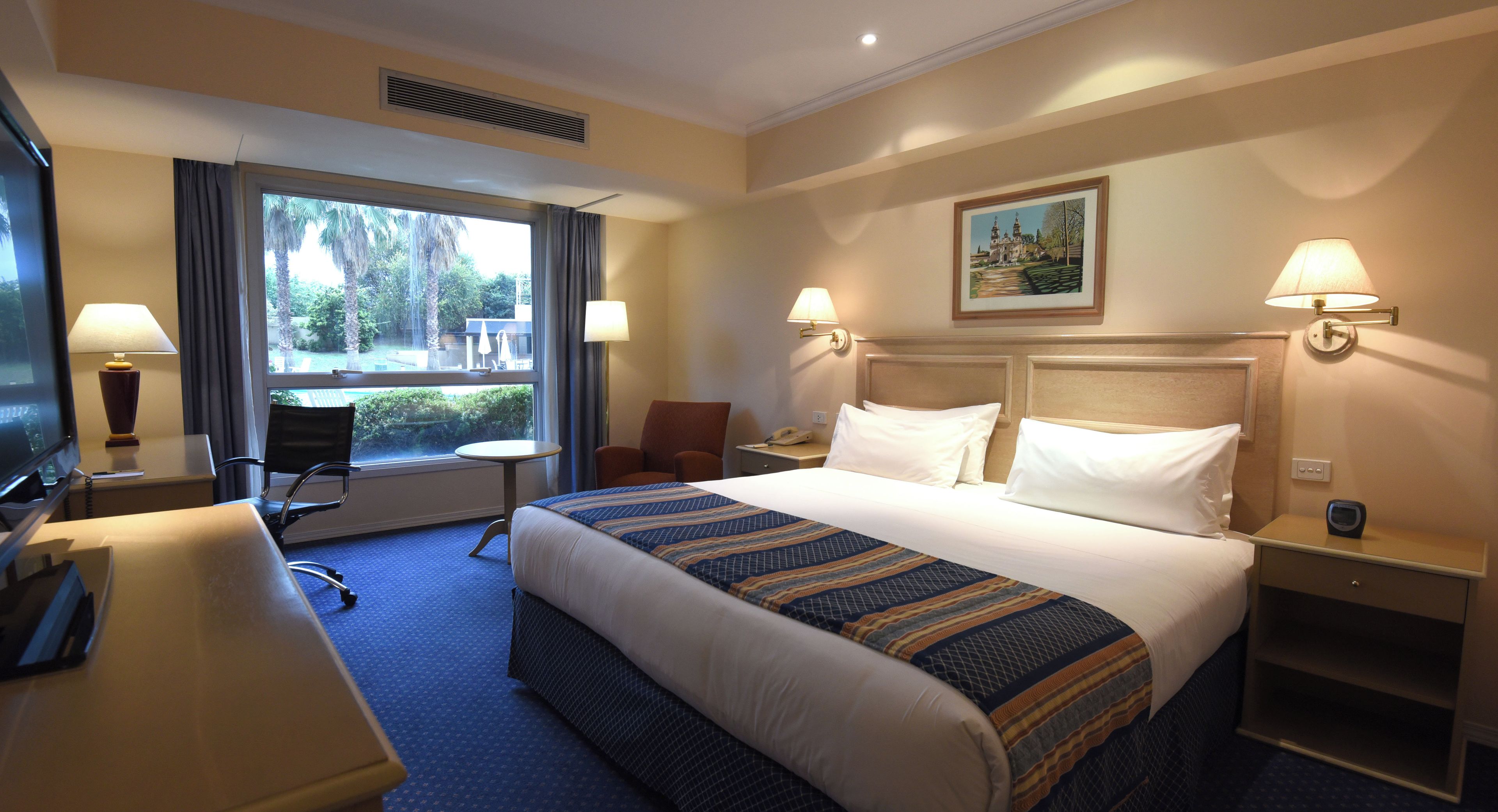Quarto de hotel com uma cama grande, duas luminárias de cabeceira, uma escrivaninha com computador, uma cadeira e uma janela com vista para palmeiras.