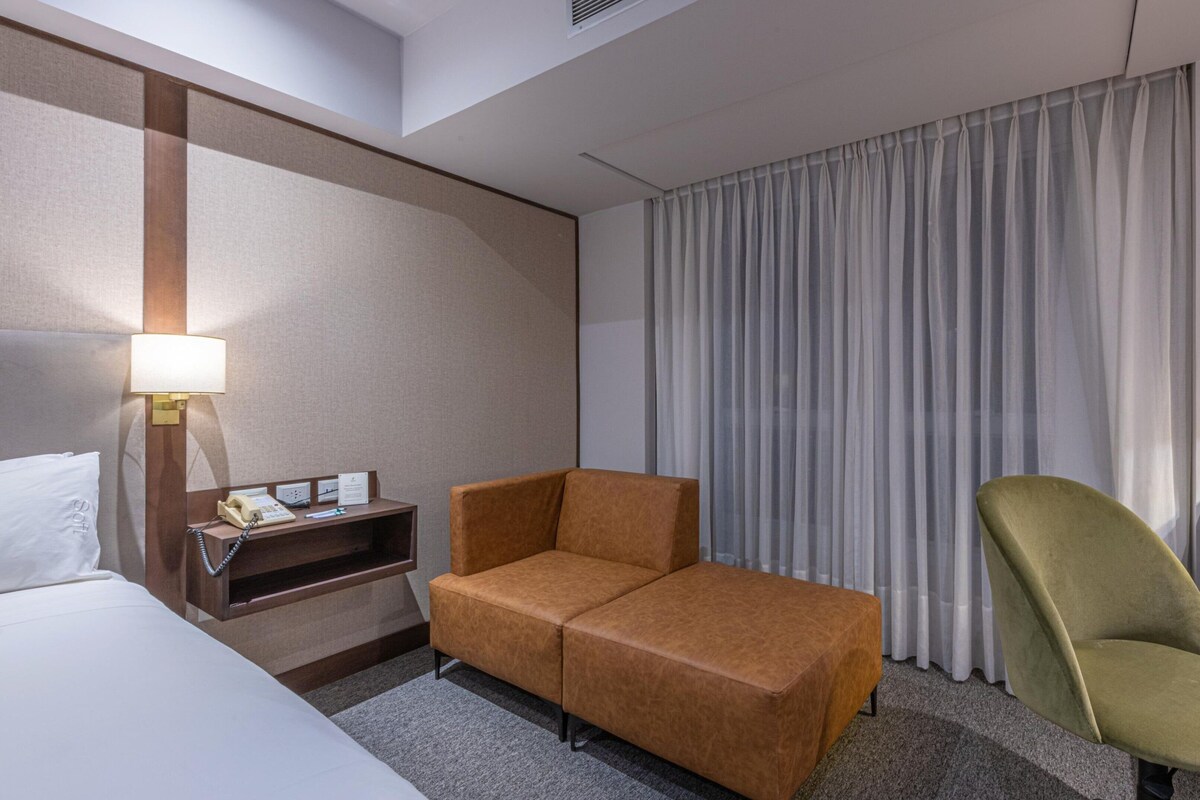 Habitación de hotel con cama, sofá, sillón, escritorio con lámpara, mesita de noche con teléfono y ventana con cortinas.