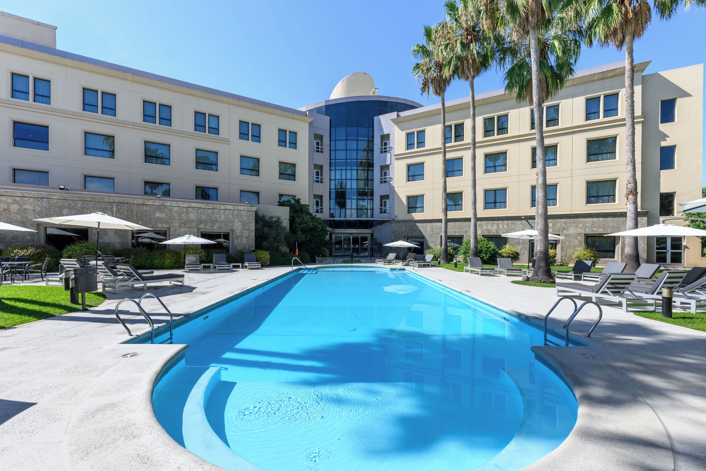 Ein Schwimmbecken mit Liegestühlen und Sonnenschirmen, umgeben von einem mehrstöckigen Hotelgebäude mit Kuppel.