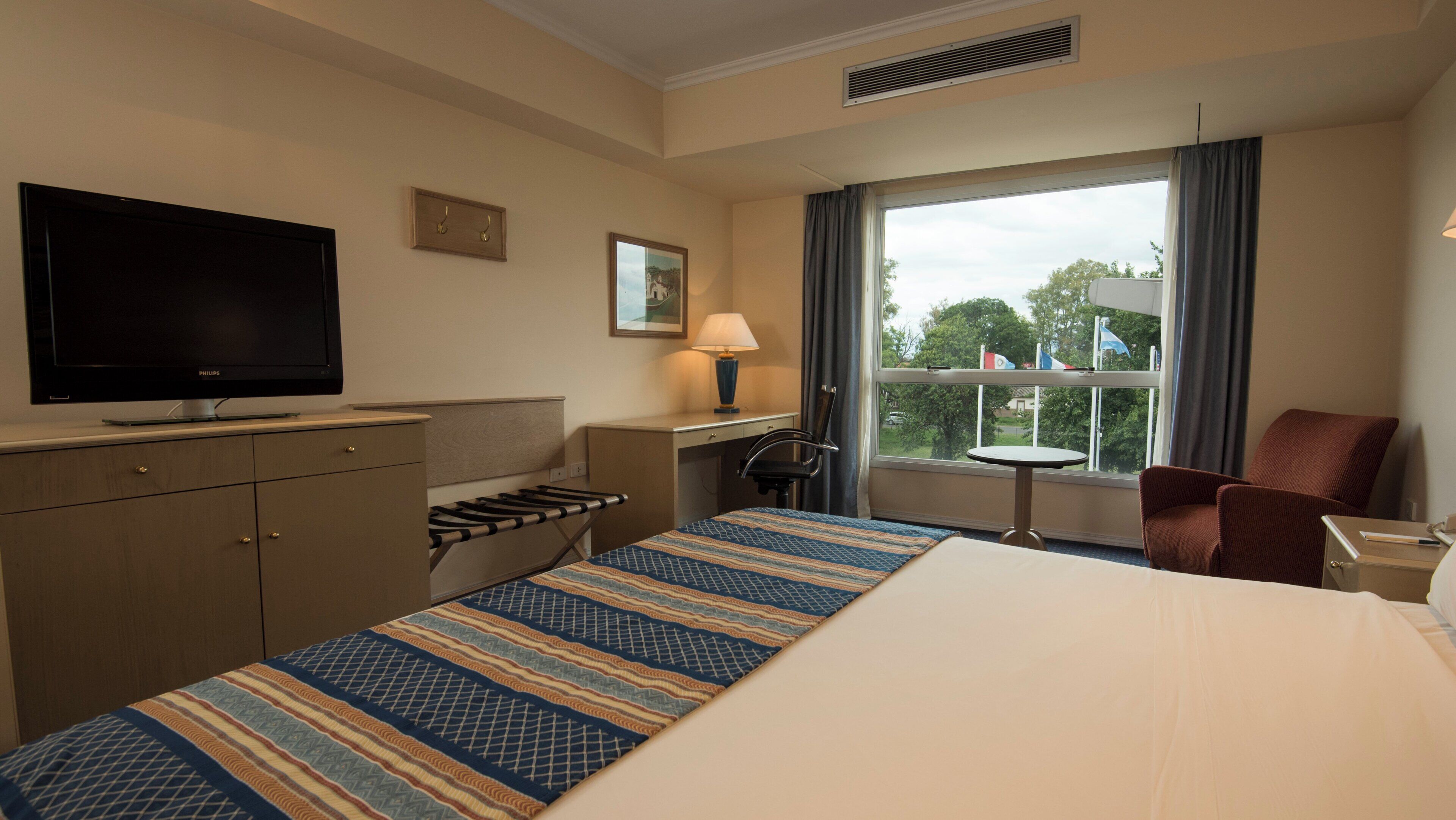 Habitación de hotel con cama, escritorio, silla, televisor y ventana con vista a árboles.