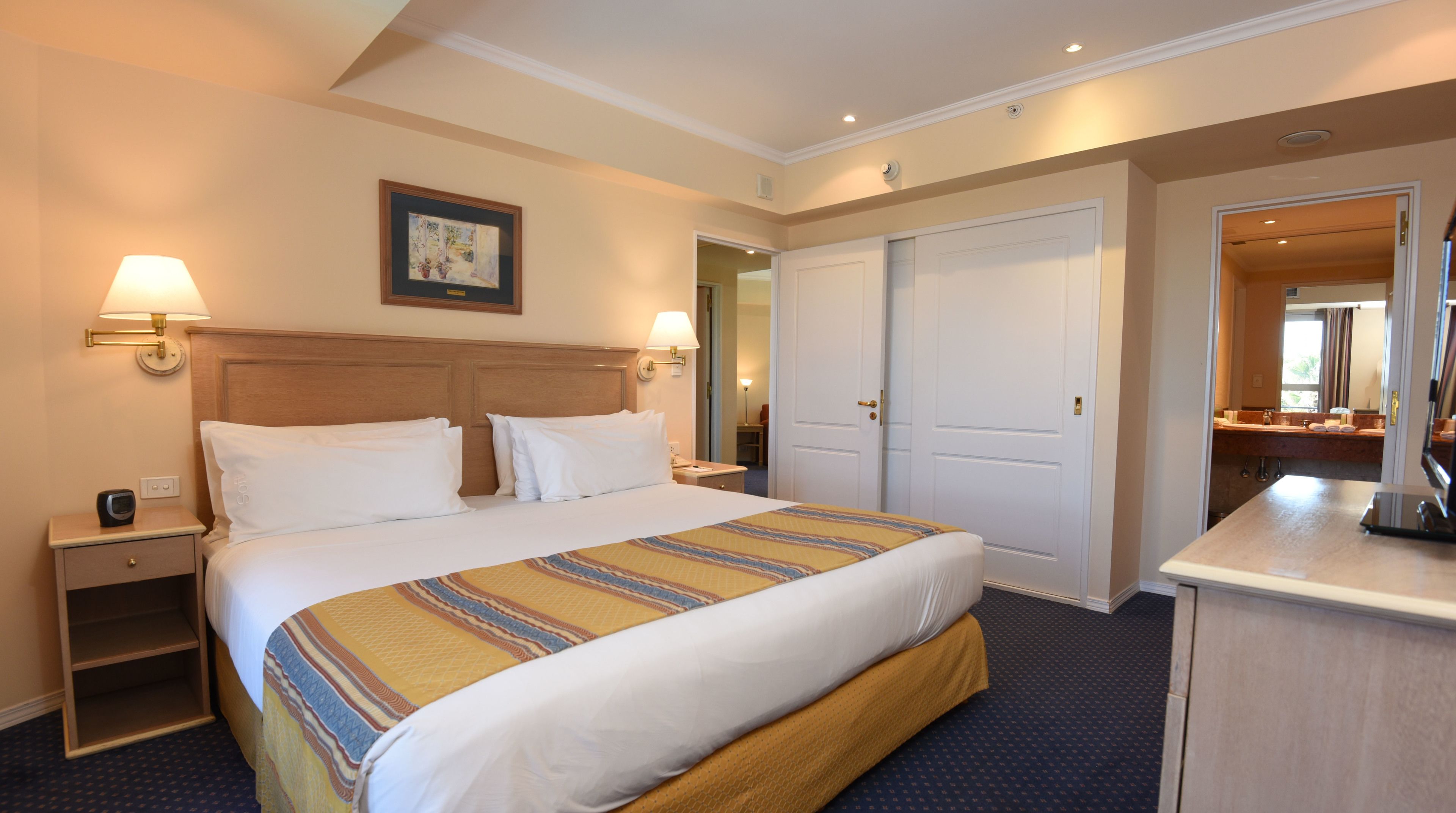 A hotel room with a bed, bedside lamps, a nightstand, a dresser, a mirror, and a door leading to another room.
