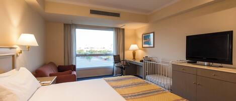 A hotel room with a bed, a desk, a chair, a television, and a window with a view of a parking lot.