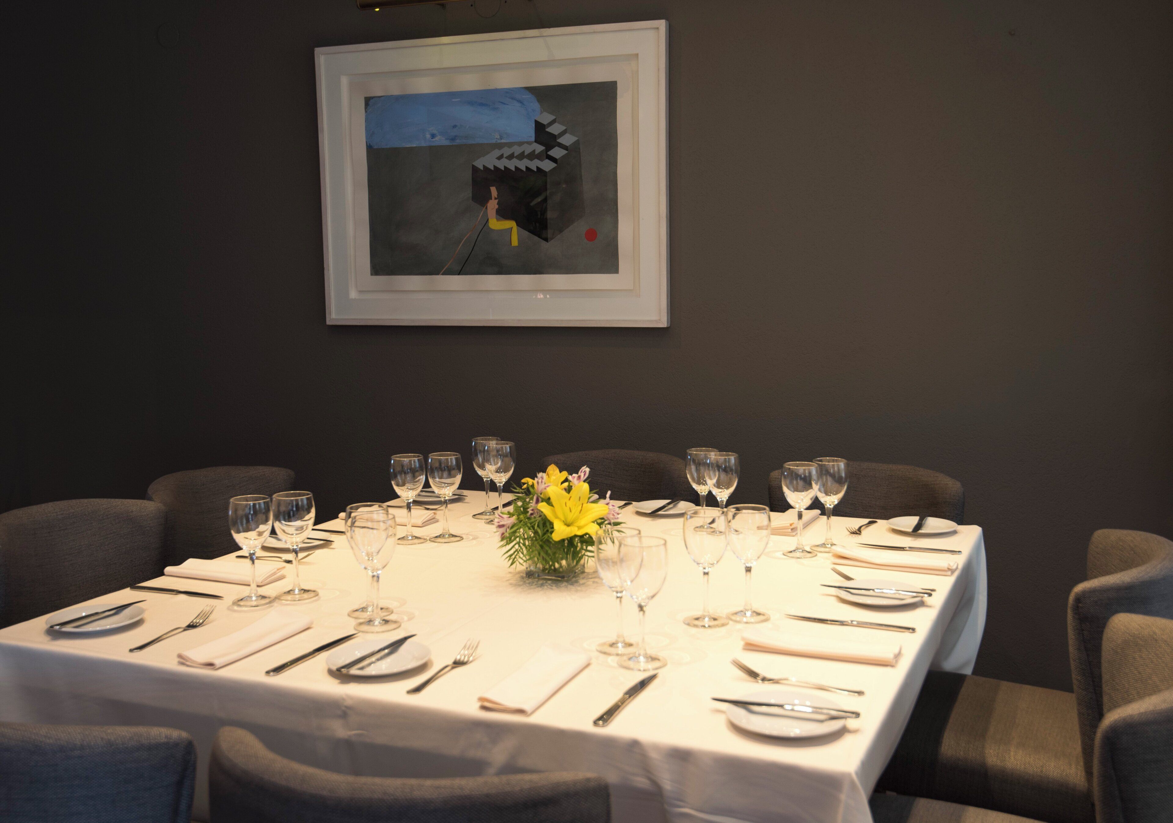 A dining table set for a meal with multiple chairs, a framed artwork on the wall, and a vase with yellow flowers.