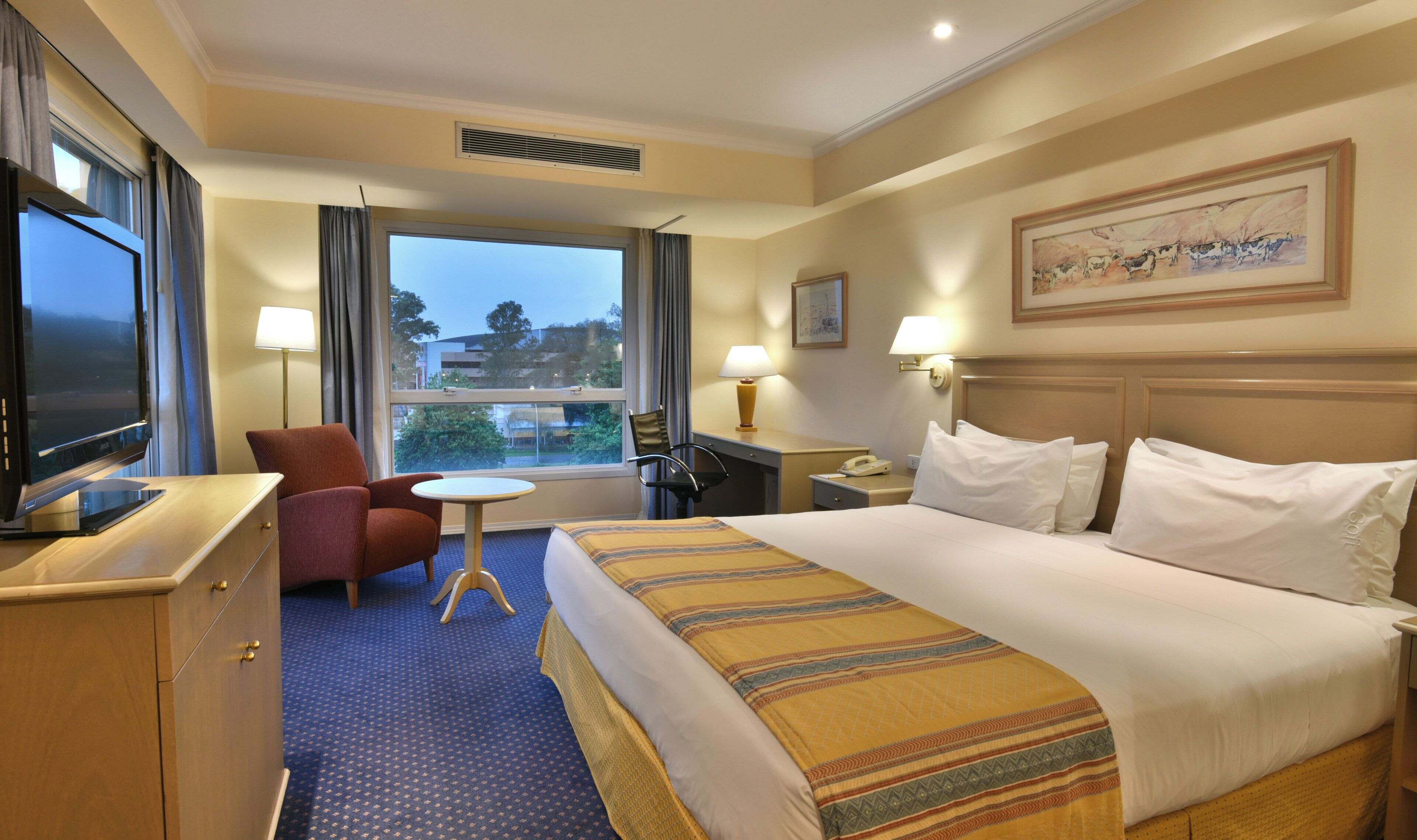 A hotel room with a bed, a desk, a chair, a television, and a window with a view of buildings and trees.
