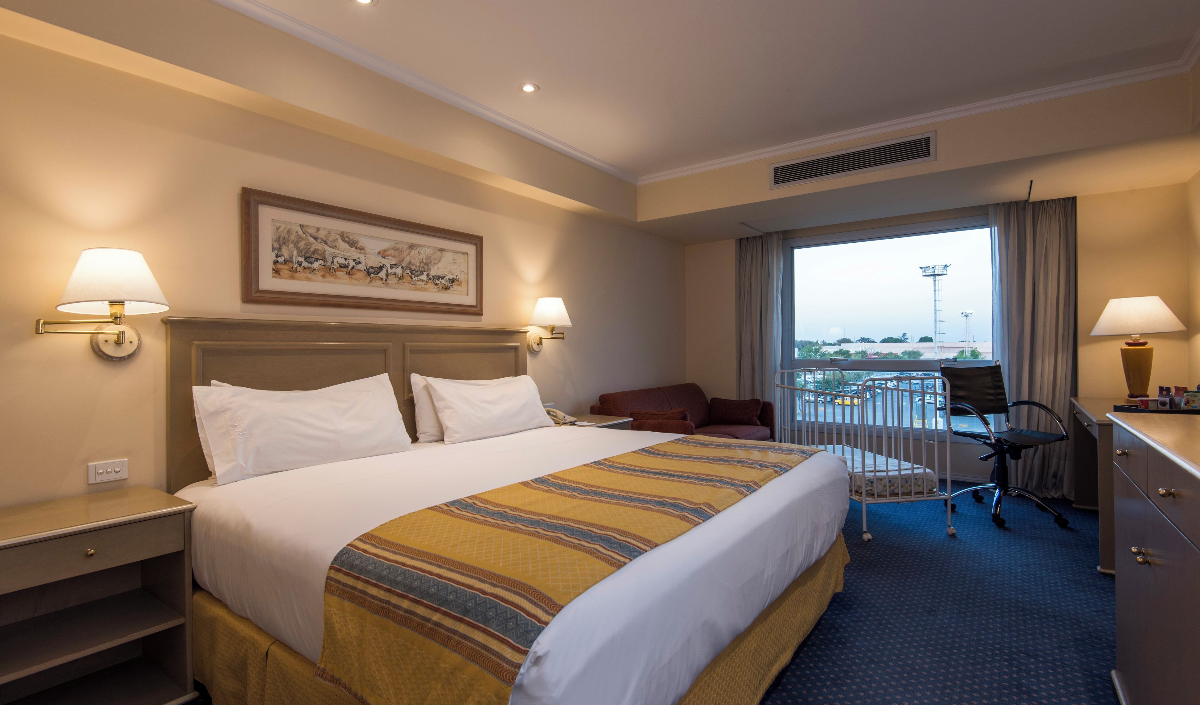 A hotel room with a large bed, two bedside lamps, a desk with a chair, a window with a view, and a painting on the wall.