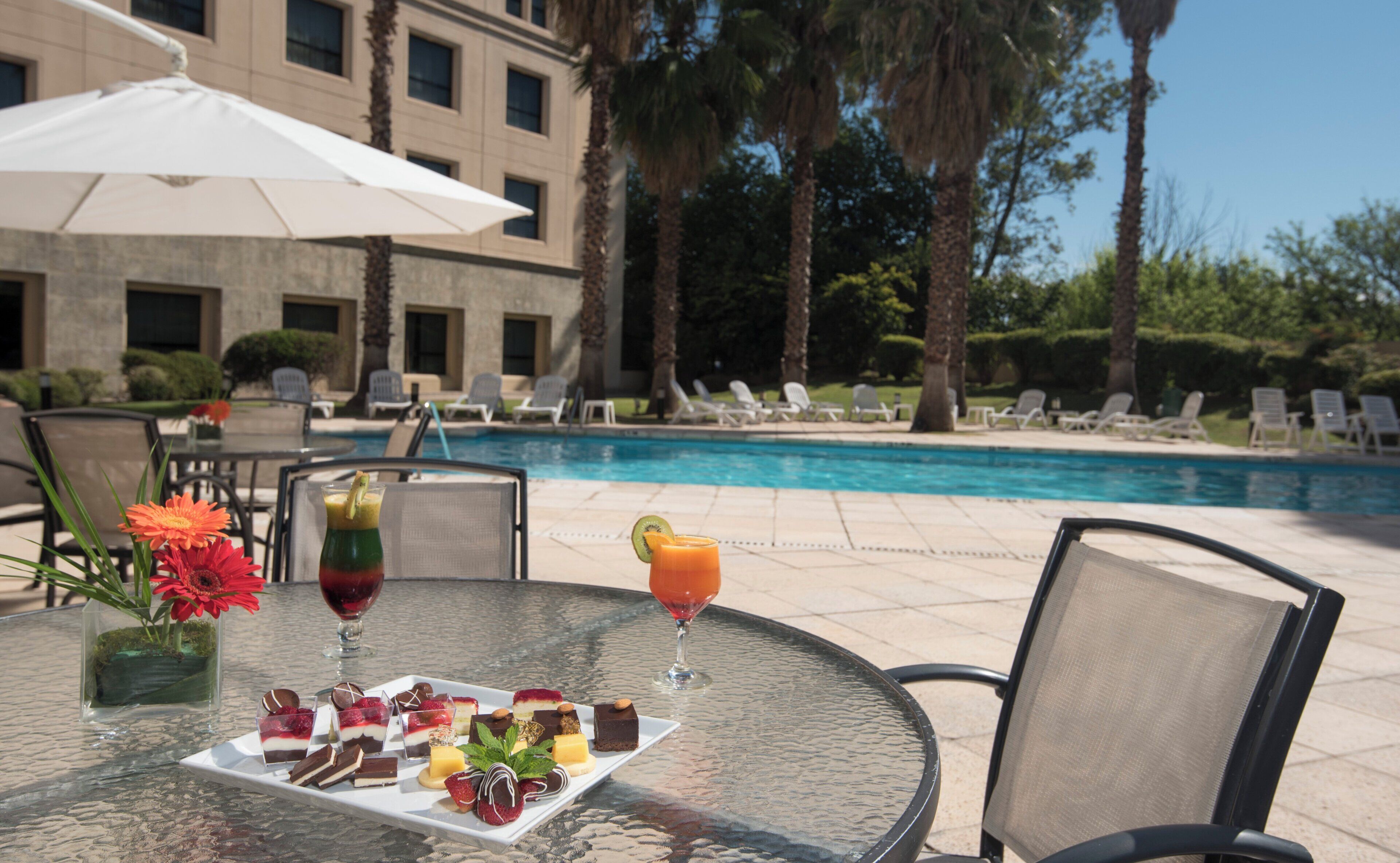 A poolside table with a dessert platter, two drinks, and a flower vase.