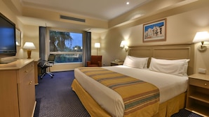 Quarto de hotel com uma cama grande, uma escrivaninha com computador, uma cadeira, uma televisão e uma janela com vista para palmeiras.