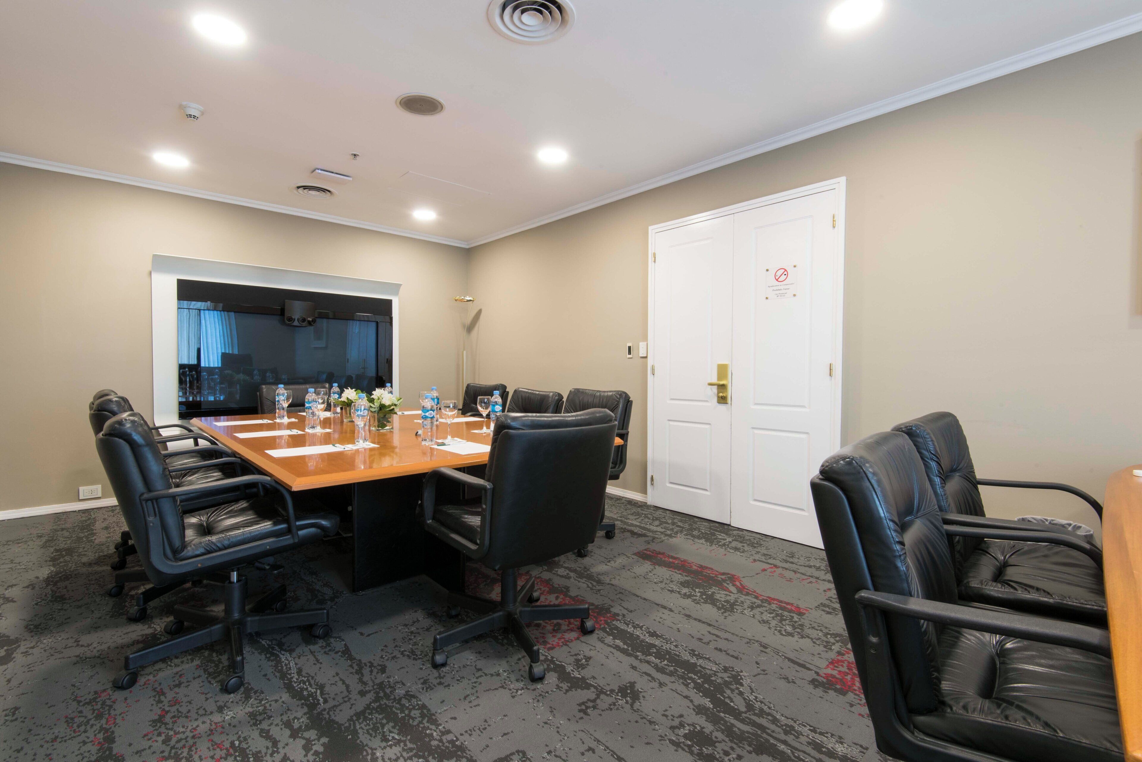 Sala de conferência com uma mesa comprida, cadeiras de couro preto e uma porta branca com placa de proibido fumar.