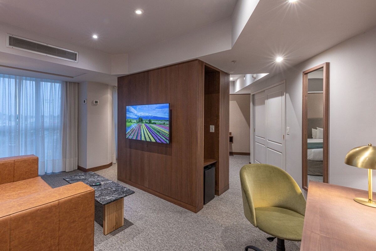 A hotel room with a large flat-screen TV mounted on a wooden panel, a brown leather sofa, a green armchair, a small wooden coffee table, and a desk with a lamp.