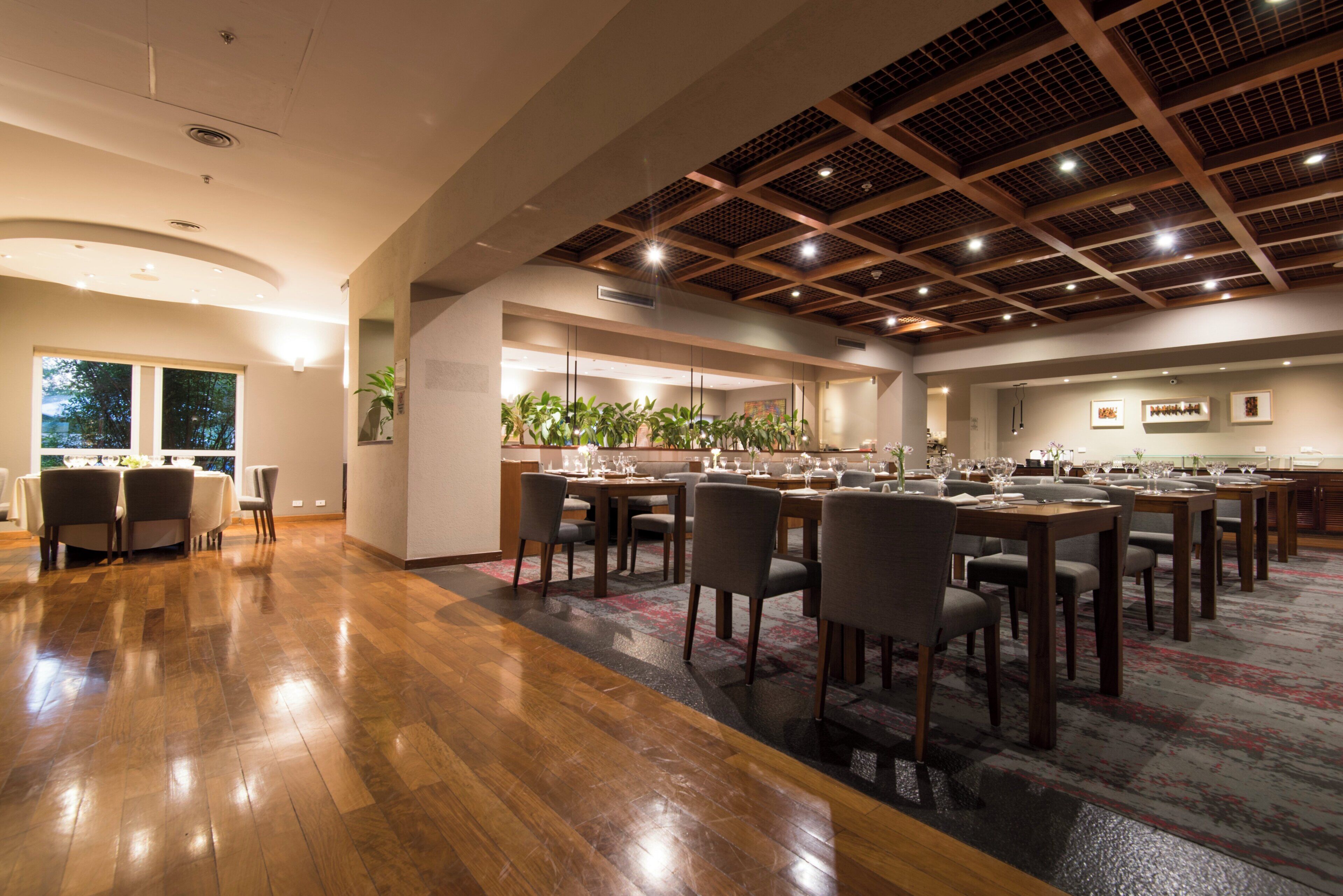A spacious dining area with multiple tables and chairs, a high ceiling with wooden beams, and a large window providing natural light.