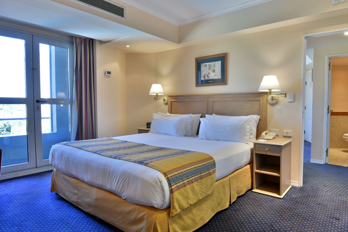 Habitación de hotel con una cama grande, mesitas de noche, un teléfono y un baño visible a través de una puerta abierta.