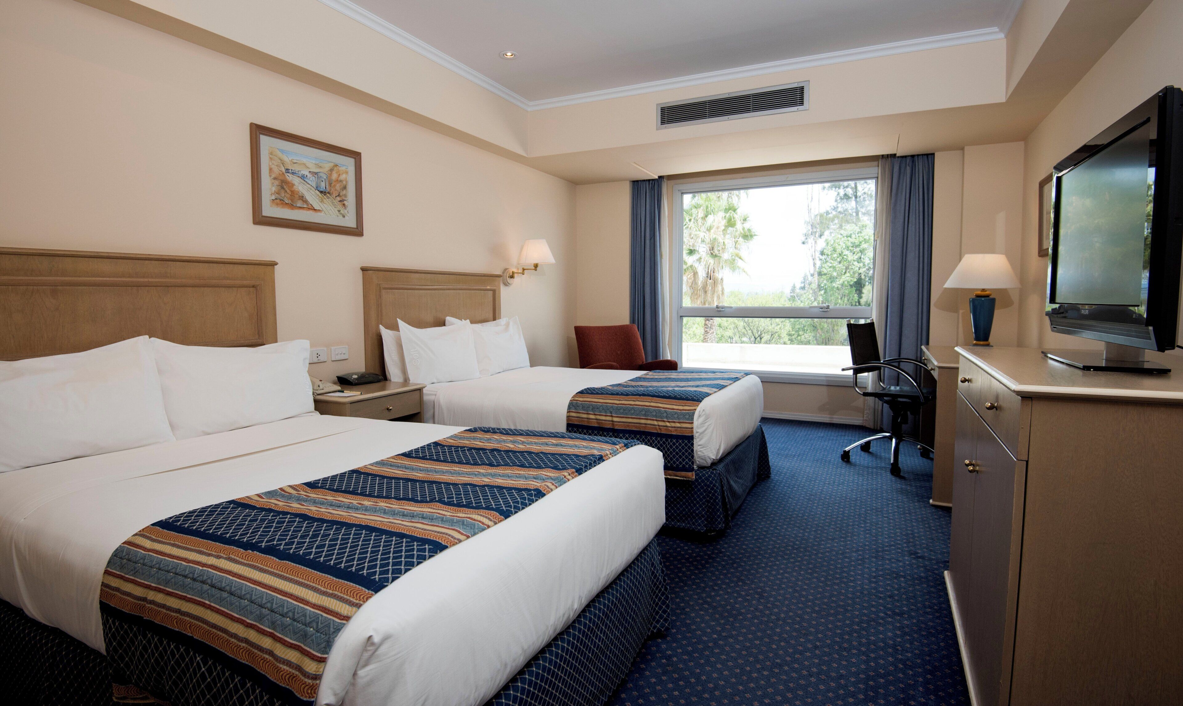 A hotel room with two beds, a desk with a television, and a window with a view of trees.