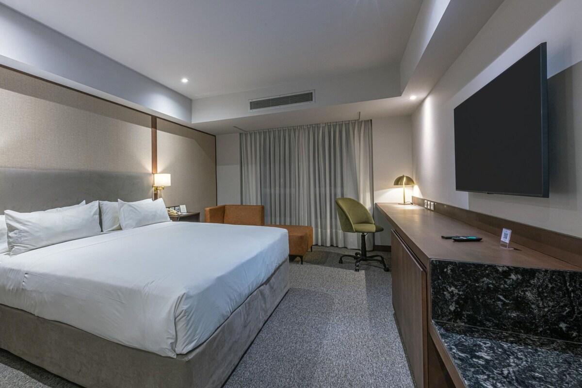 Habitación de hotel con una cama grande, un escritorio, una silla, un televisor de pantalla plana y un ventanal con cortinas.