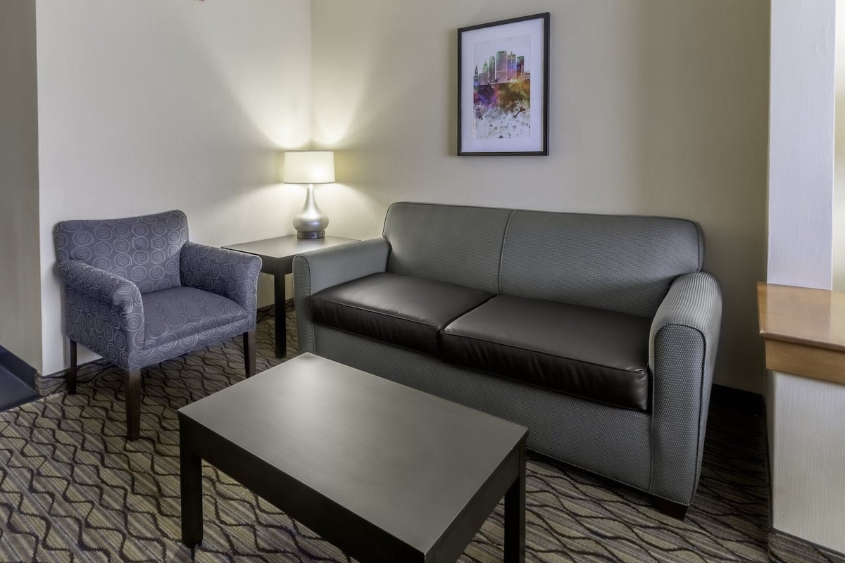 Hotellihuone, jossa on harmaa, kuviollinen nojatuoli, harmaa sohva, pieni pöytä ja seinällä kehystetty kaupunkimaisemakuva.