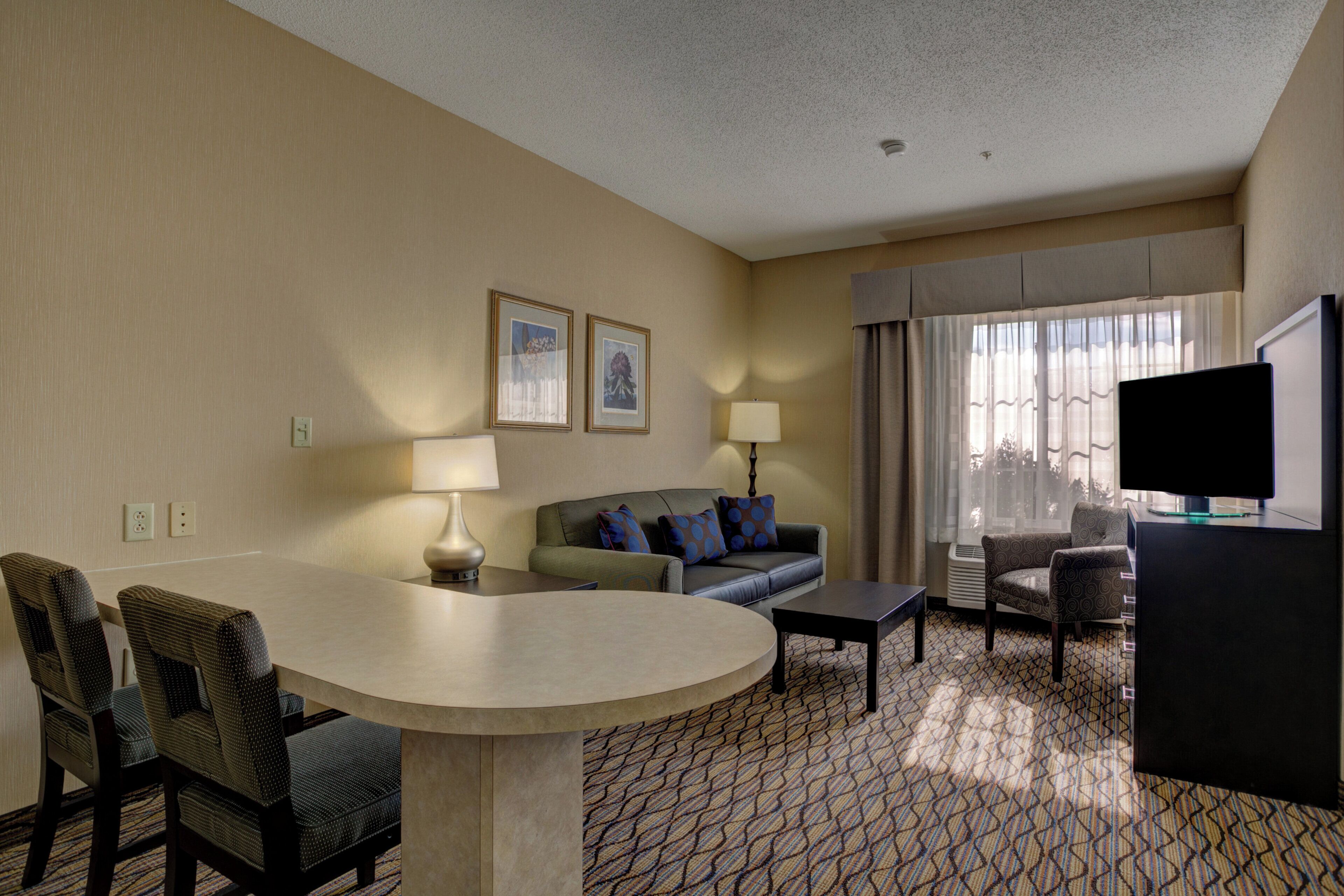 A hotel room with a curved desk, two chairs, a sofa with patterned cushions, a television, and a window with curtains.