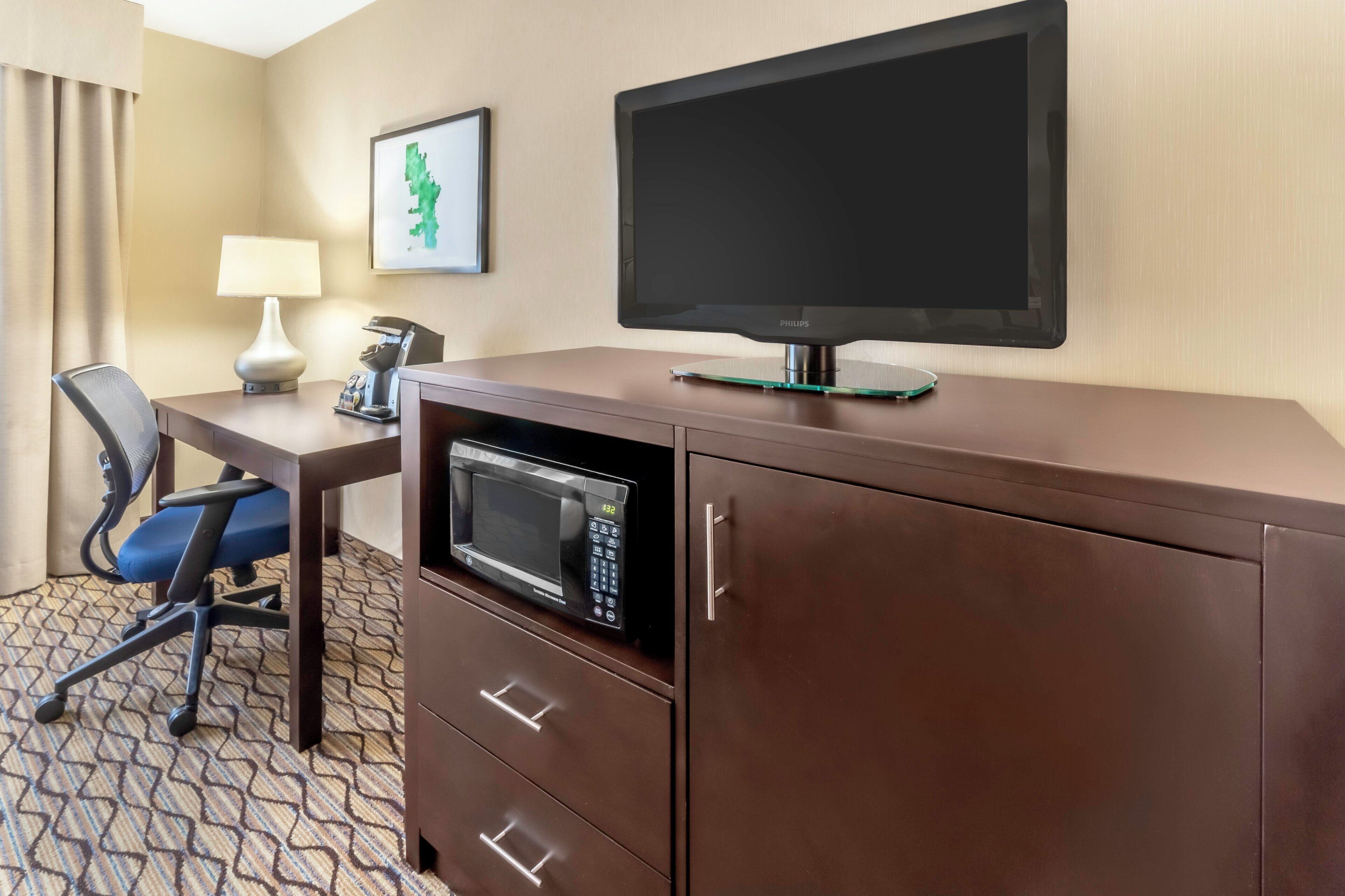 A hotel room with a flat-screen TV, a microwave, a desk with a lamp, and a chair.