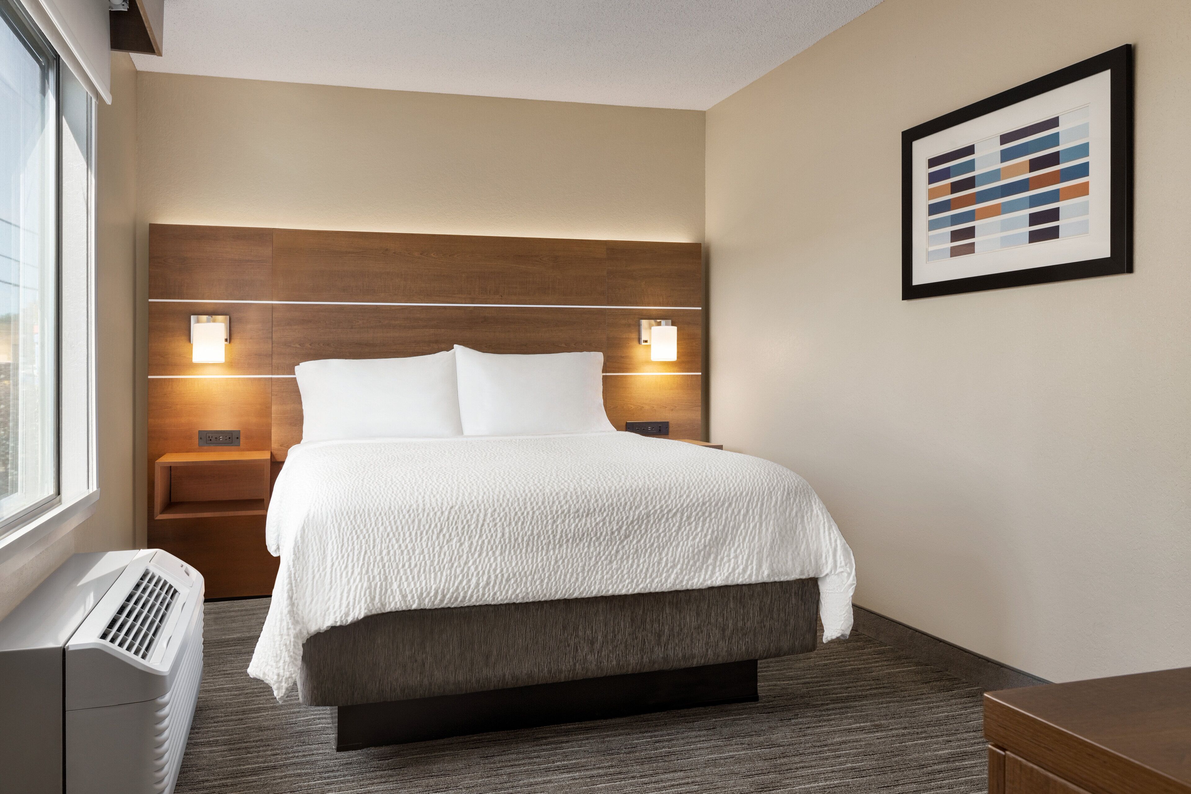 A hotel room with a large bed, two bedside tables, a wall-mounted light fixture, a framed picture, and a window.