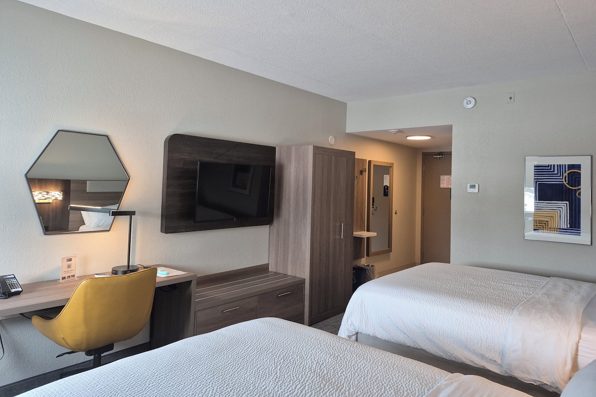 A hotel room with two beds, a desk with a chair, a mirror, a TV, and a door leading to a bathroom.