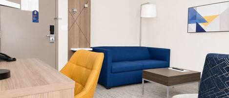 Habitación de hotel con escritorio de madera, sofá azul, silla amarilla, sillón estampado, una mesita, lámpara y un cuadro en la pared.