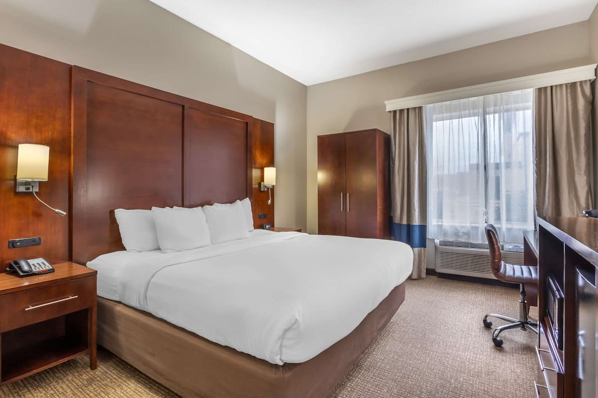 Habitación de hotel con una cama grande, cabecero de madera, lámparas de noche, un escritorio con teléfono, una silla y un ventanal con cortinas.