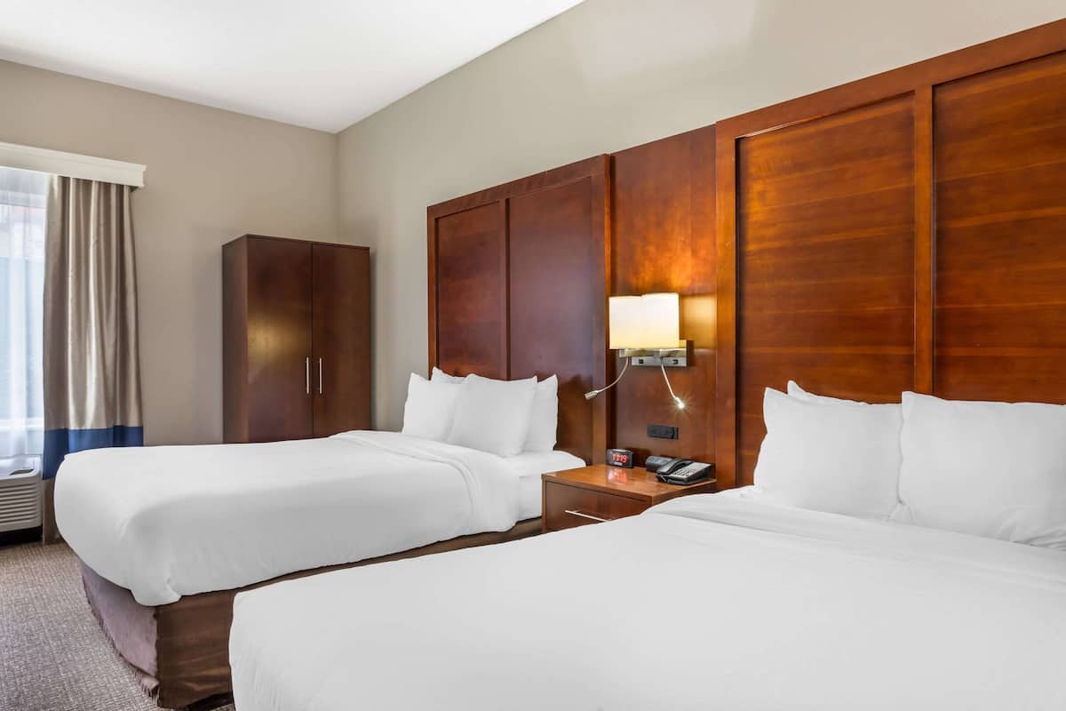 Habitación de hotel con dos camas, un armario de madera, una mesita de noche con lámpara y una ventana con cortinas.