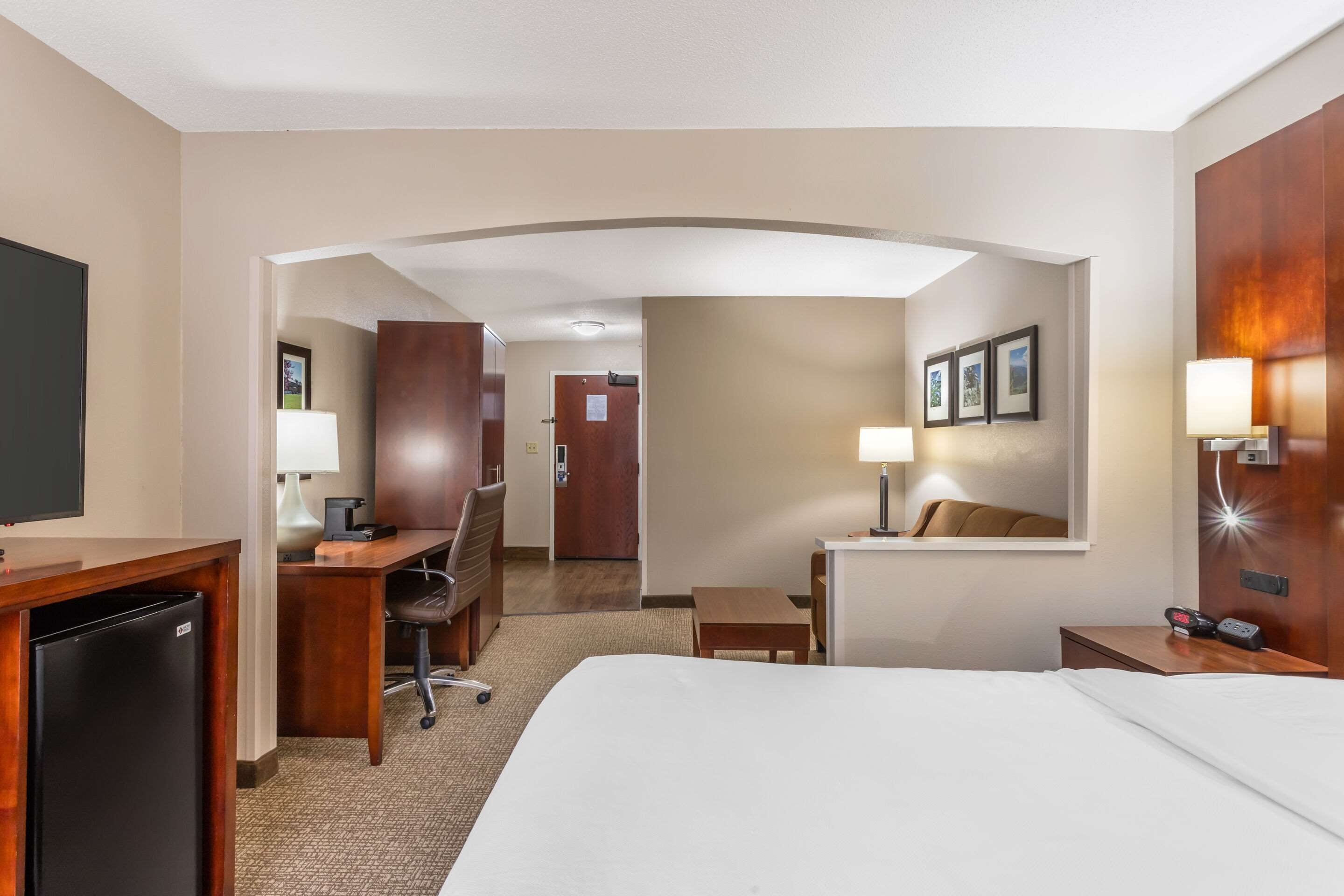 A hotel room with a large bed, a desk with a chair, a small table, a TV, and a desk lamp.