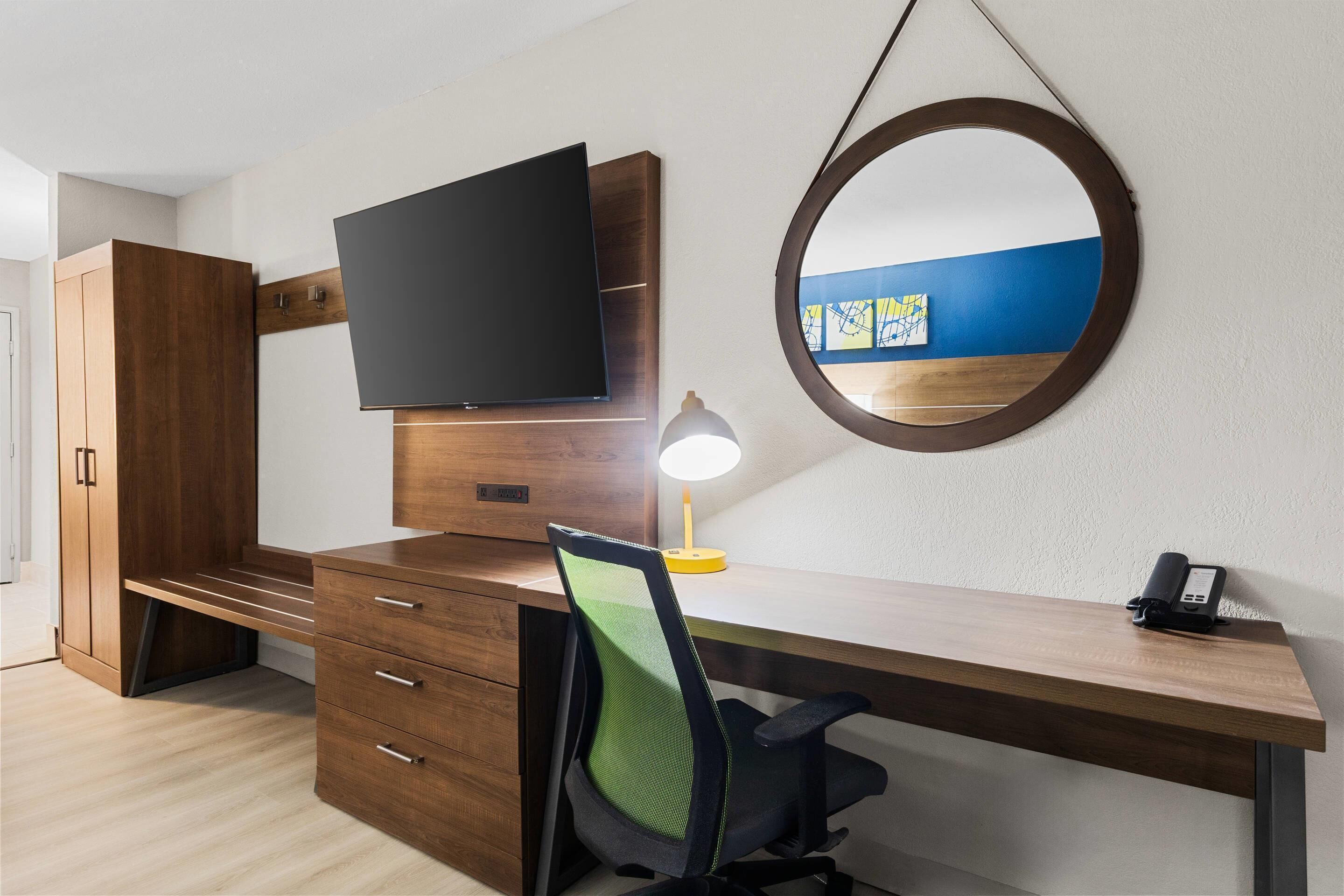 Une chambre moderne avec un bureau en bois, un téléviseur à écran plat, une lampe de bureau, une chaise, un téléphone et un miroir rond accroché au mur.