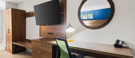 Une chambre moderne avec un bureau en bois, un téléviseur à écran plat, une lampe de bureau, une chaise, un téléphone et un miroir rond accroché au mur.