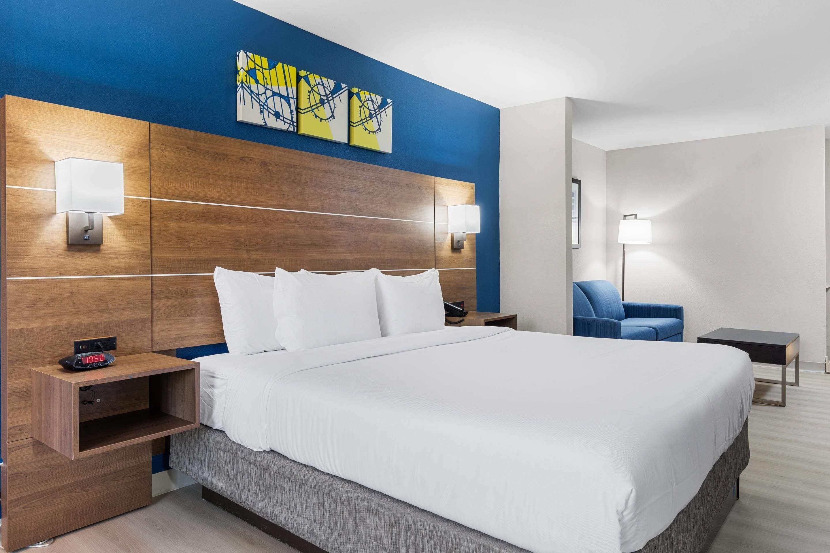 A hotel room with a large bed, wooden headboard, two bedside tables with lamps, a blue chair, and artwork on the wall.