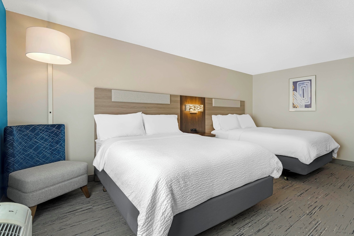 A hotel room with two beds, a desk, a chair, a lamp, and a wall-mounted light fixture.