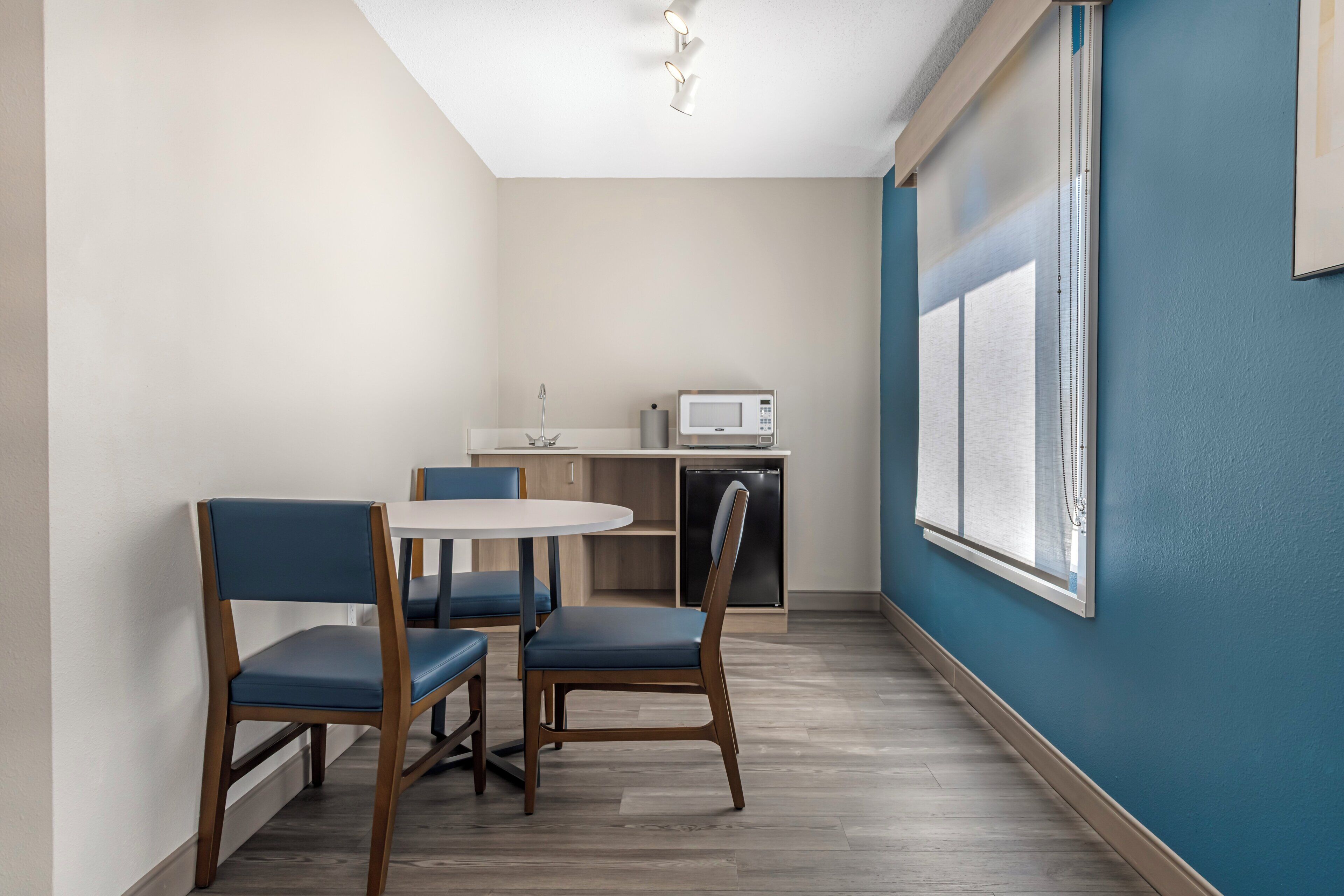 A dining area with a round table and four chairs, a microwave, and a refrigerator in the background.