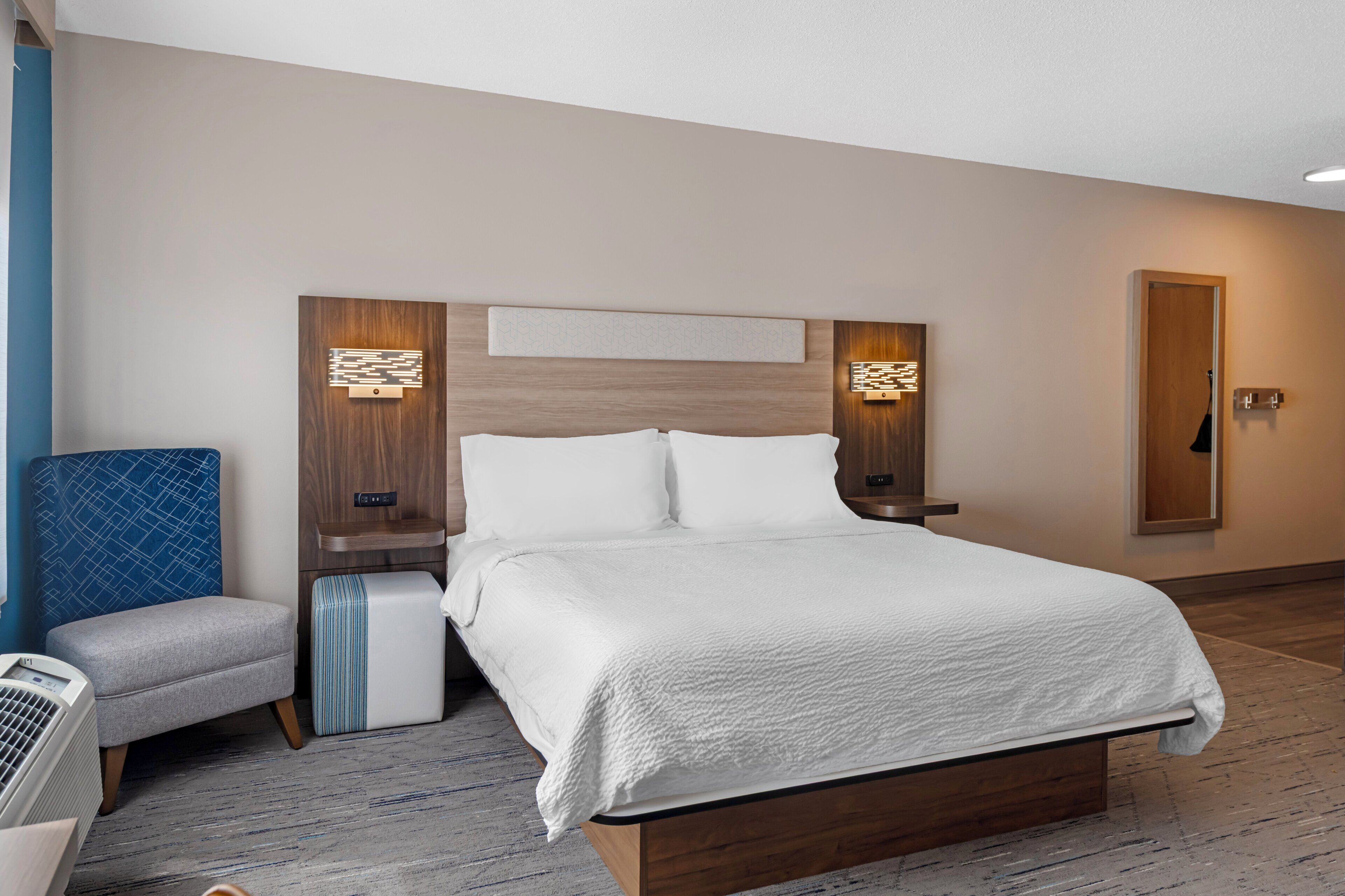 A hotel room with a bed, bedside tables, a chair, a small table, a mirror, and a wall-mounted air conditioner.